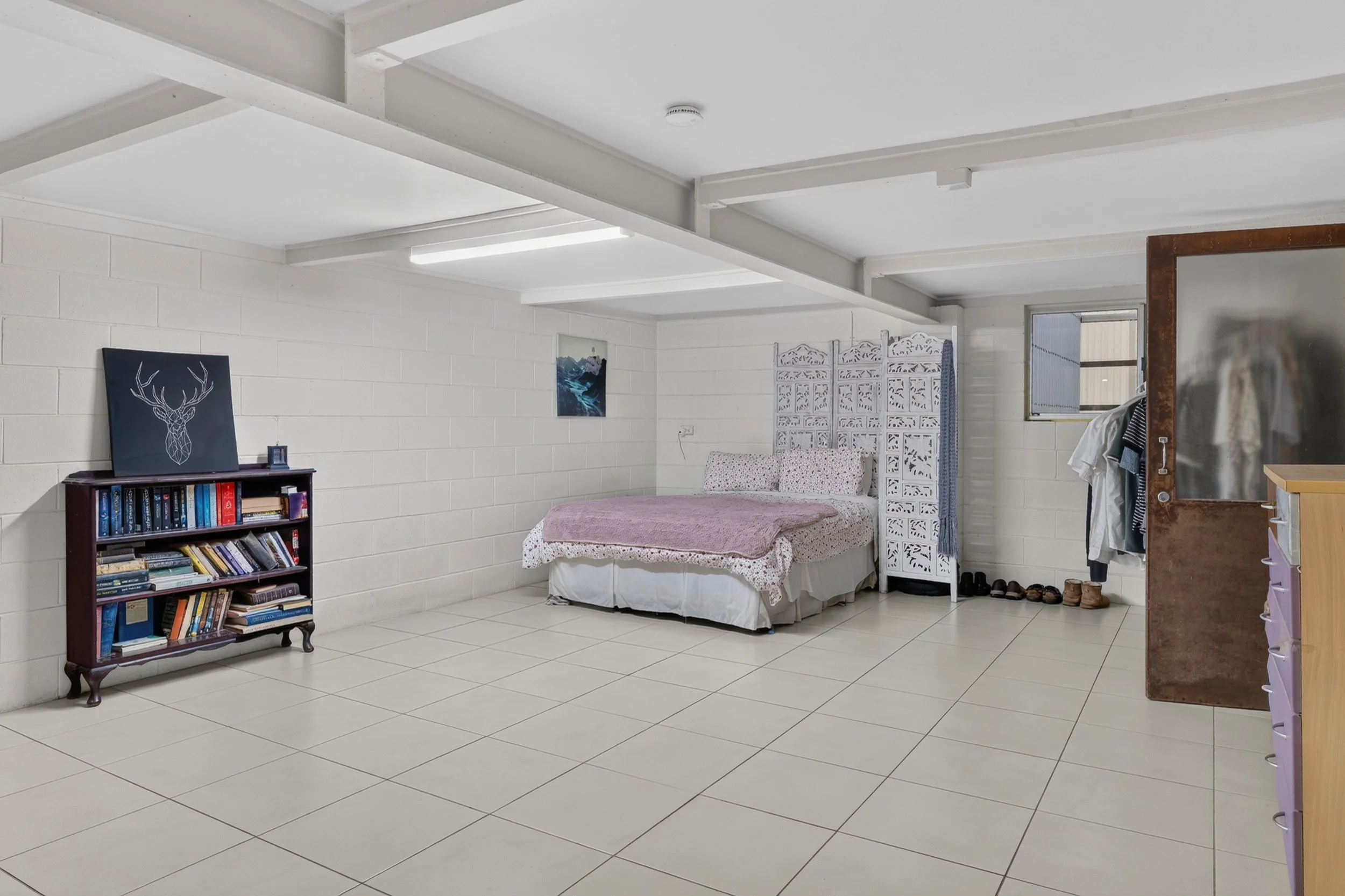A bedroom with white brick walls, white tile floor, and white ceiling. There is a bed with a pink blanket and pillows against the back wall. To the left, a bookshelf holds books and a black artwork with a white geometric deer head. There is a small b