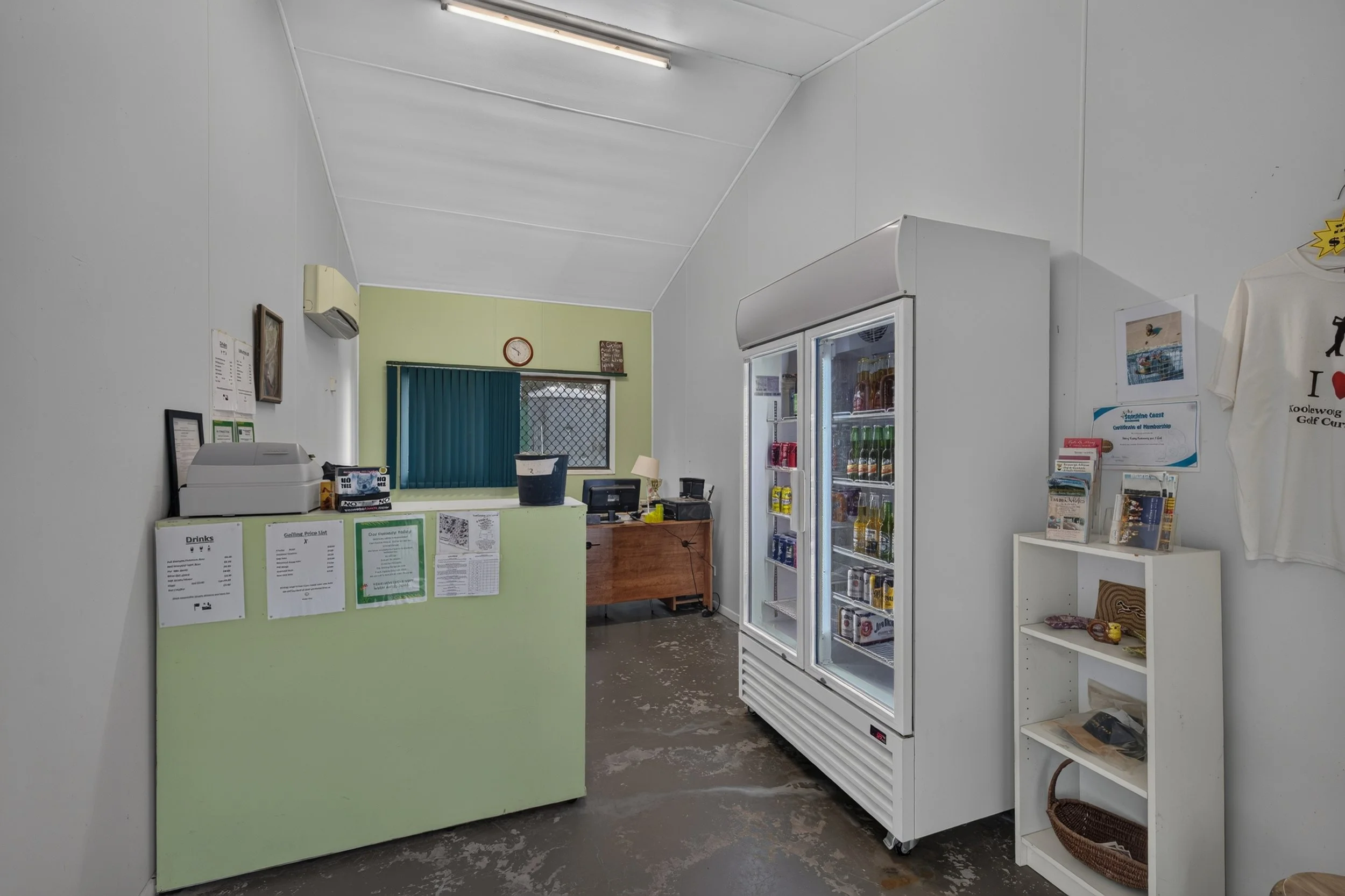 Interior of a small store or shop with a light green counter, a glass-door beverage refrigerator, and shelves with products. The store has white walls and a gray concrete floor. There is a clock, some pictures, and papers on the wall behind the count