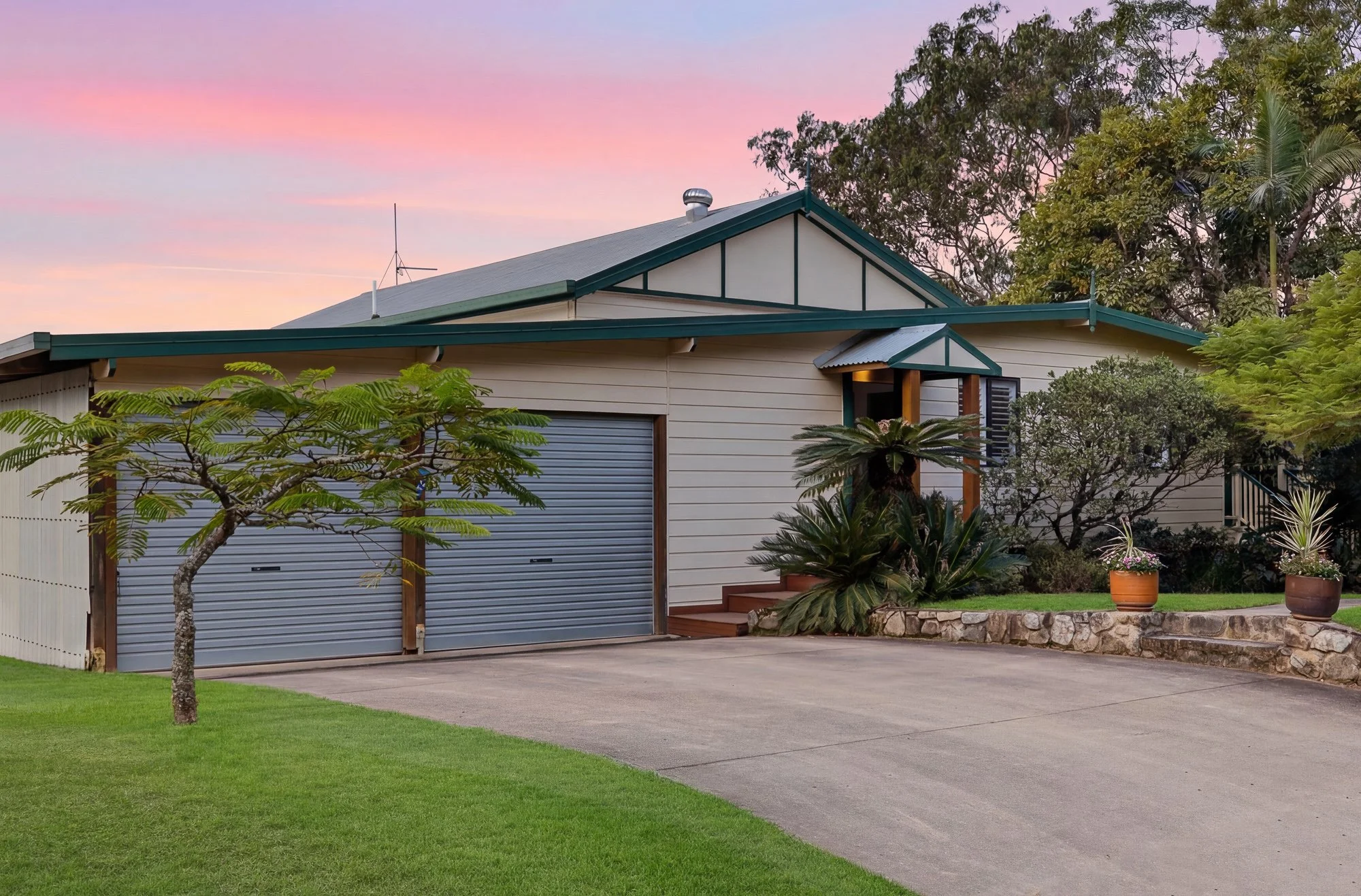A modern house with a green roof and beige siding, surrounded by lush plants and trees, with a pink and purple sunset sky in the background.
