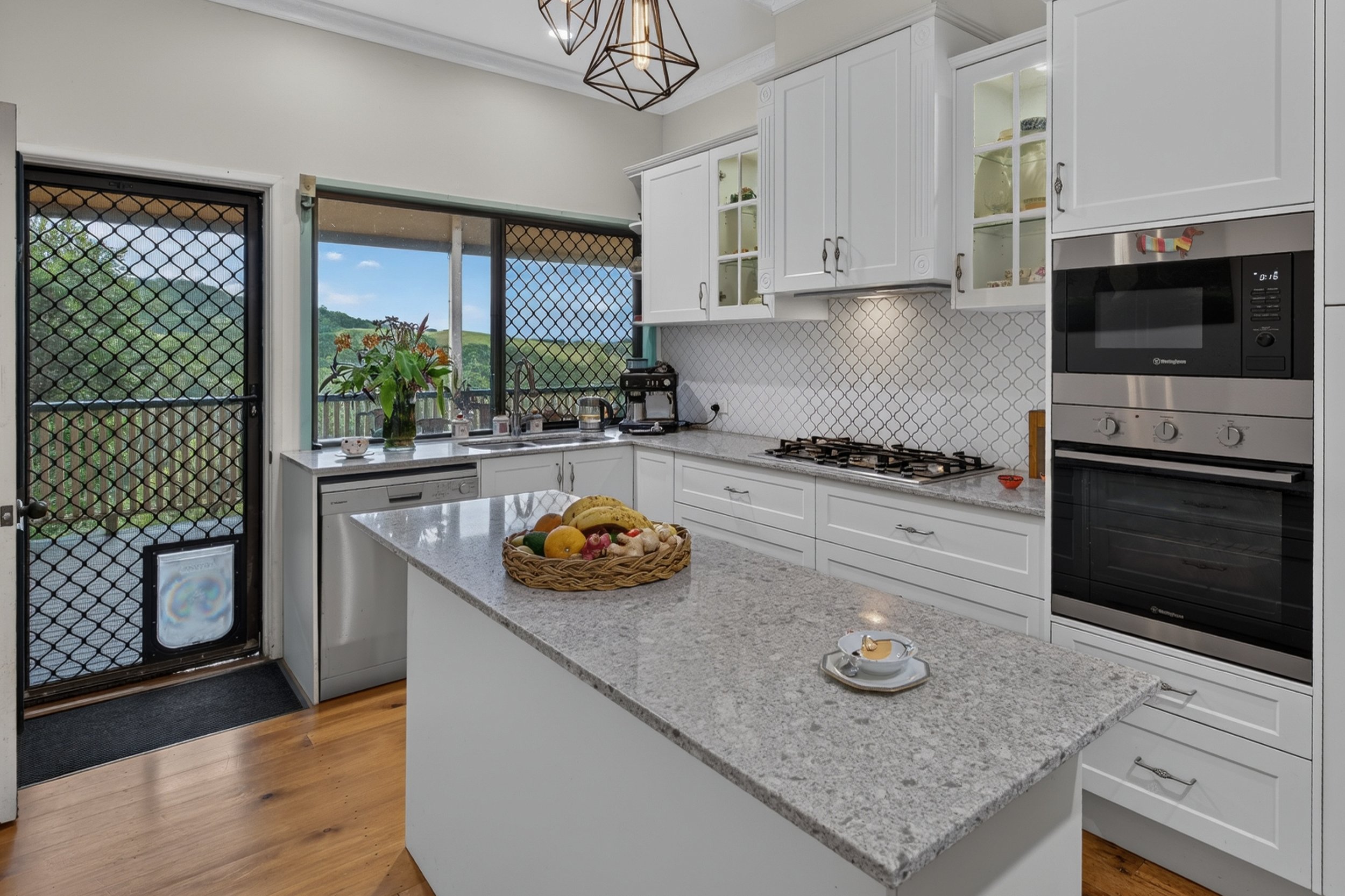 A modern kitchen with white cabinets, a gray granite countertop, and a basket of assorted fresh fruits on the island. Large windows and a glass door with a security screen provide a view of lush green hills outside. The kitchen features built-in appl