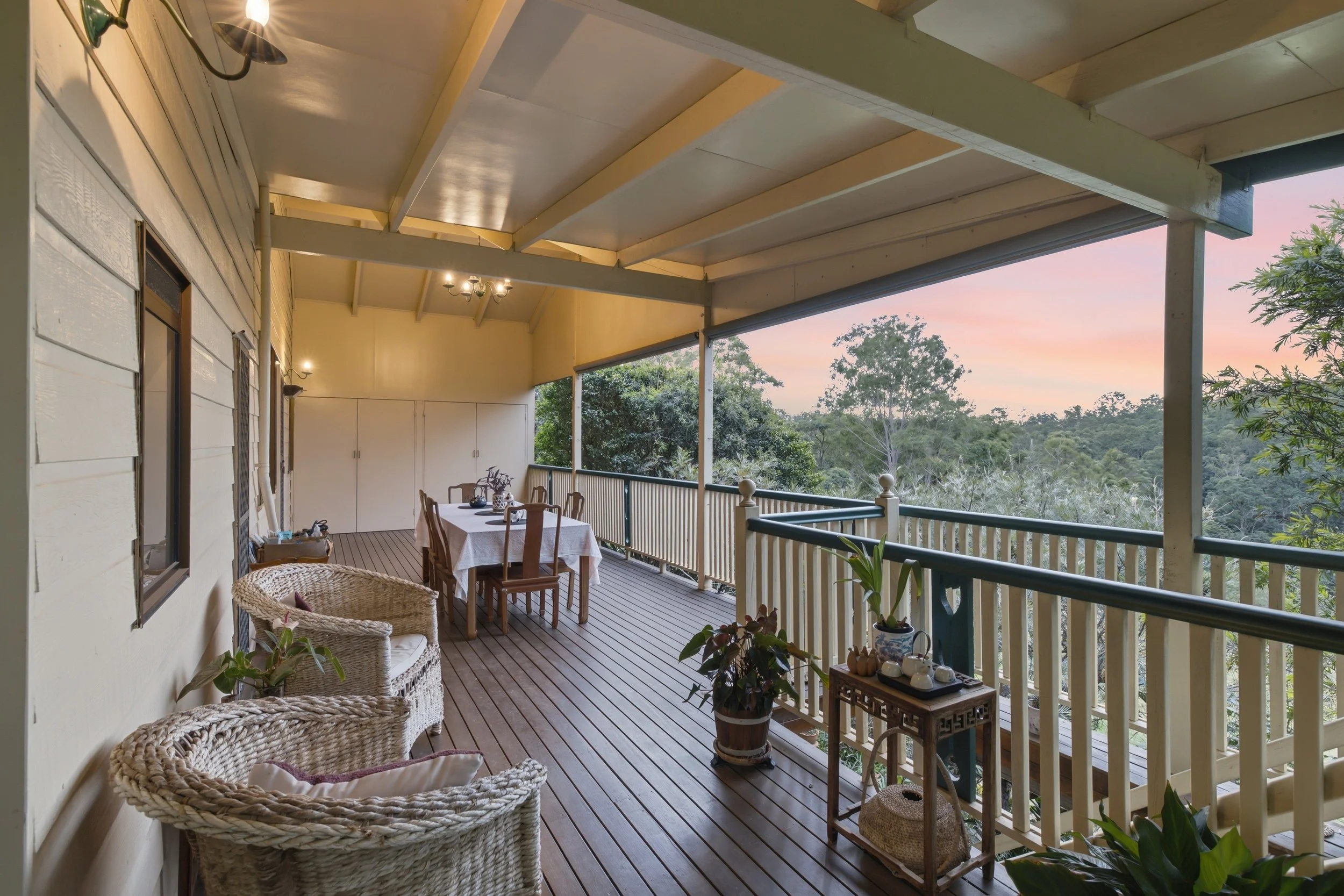 A spacious covered porch with a wooden floor, beige walls, and a railing overlooking a lush green landscape. There is a round dining table with a white tablecloth and chairs, wicker seating, various potted plants, wall-mounted lamps, and a storage ca