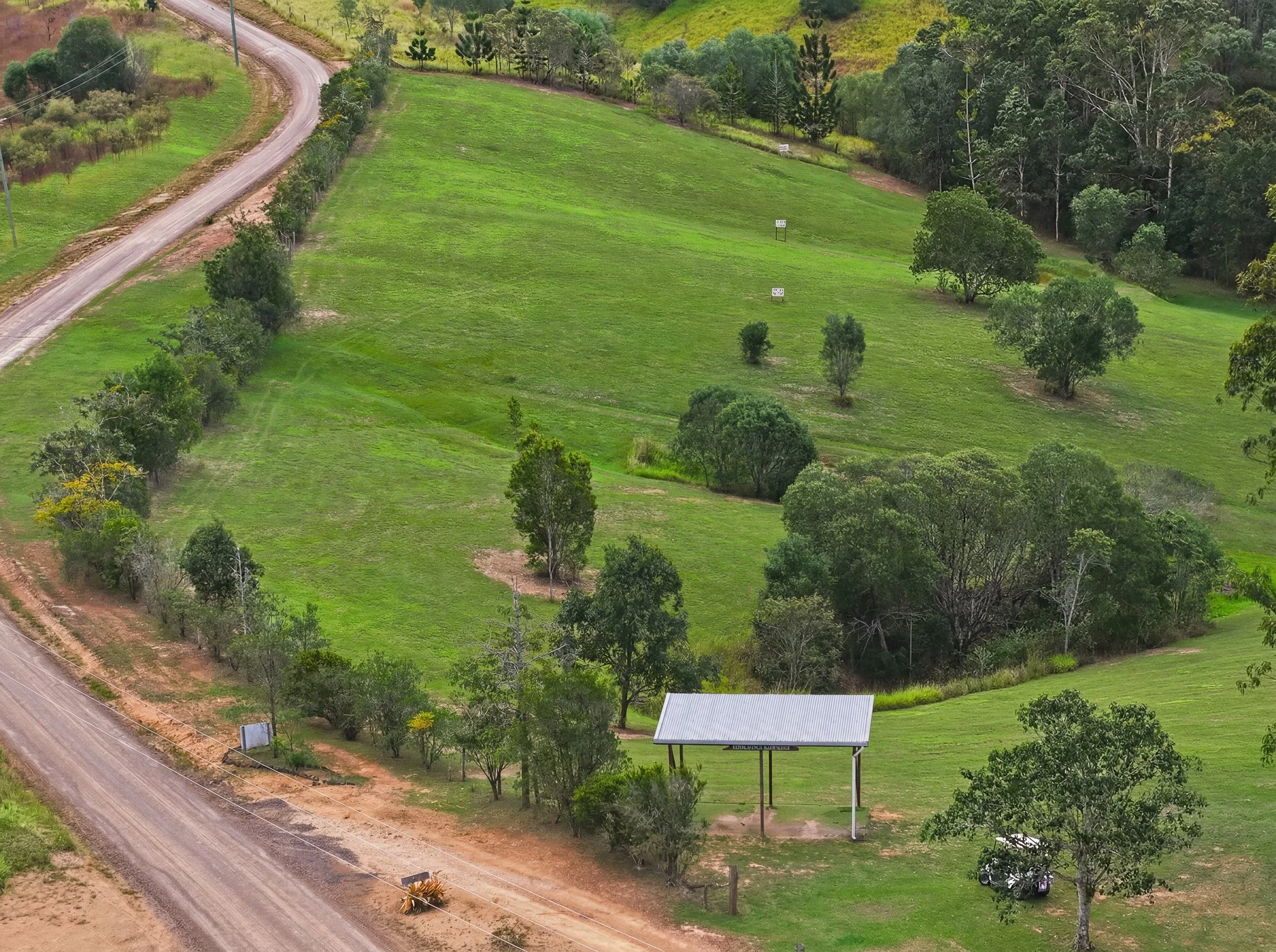A landscape view of a green hillside with a winding dirt road on the left side, lined with various trees and shrubs. A small shelter with a sign is visible near the bottom right corner of the image.