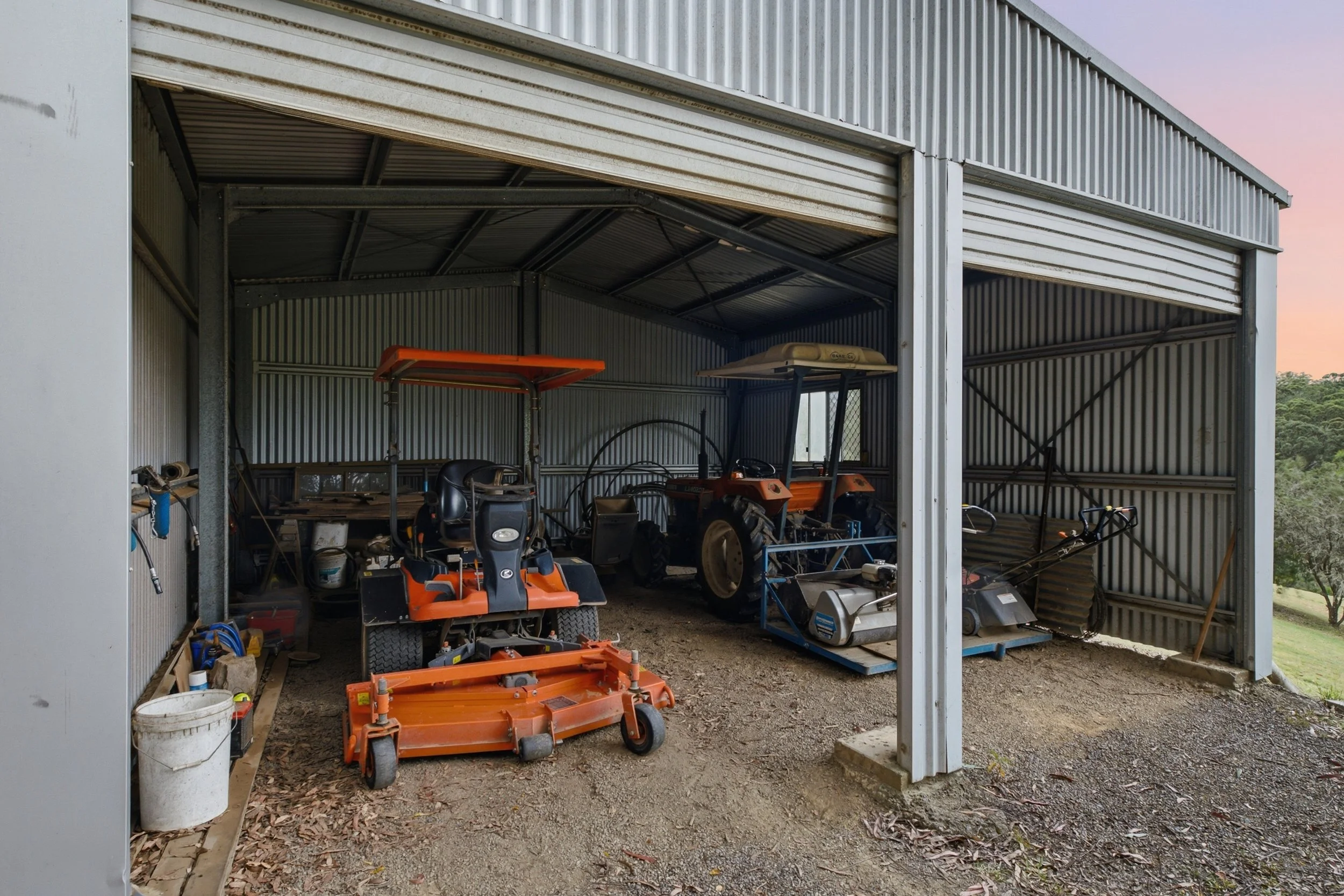 Inside view of a metal shed with farming equipment and tools, including a lawn mower, a small tractor, and various other items.