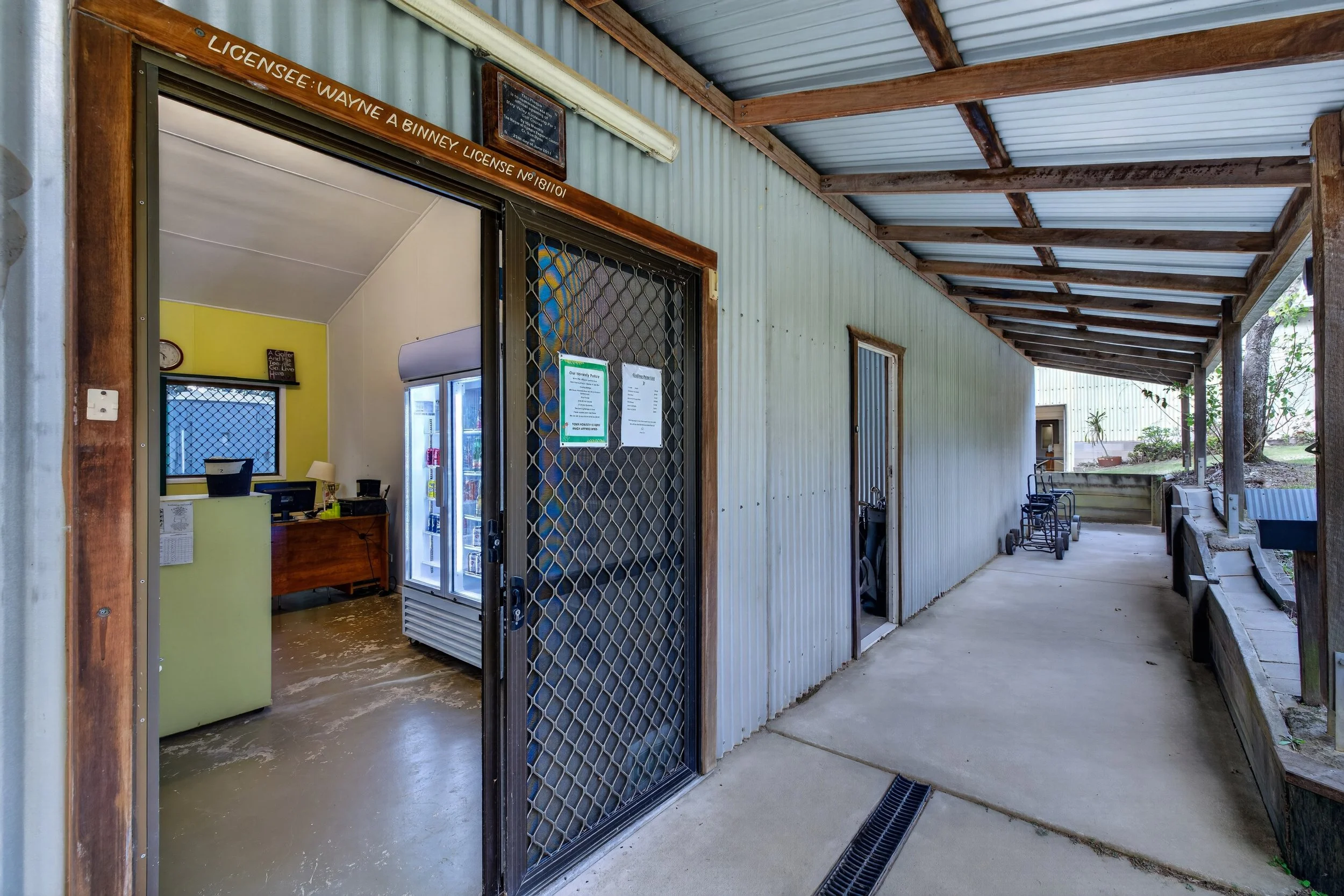 View of an outdoor covered patio area with a concrete floor, a wooden ceiling with exposed beams, and metal siding walls. There are chairs, a fish tank, windows, and various signs and notices on the wall.