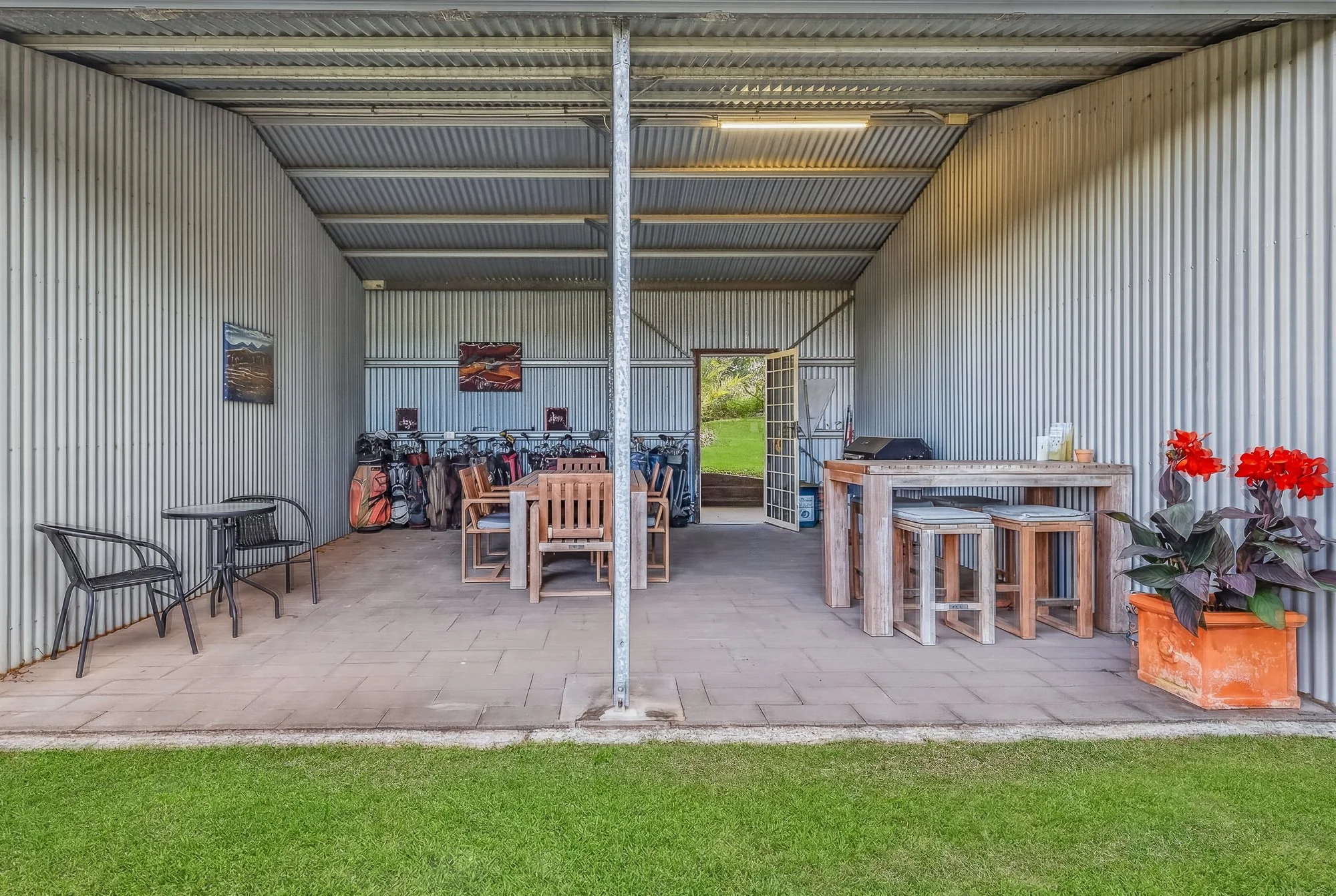 Backyard patio with metal walls and ceiling, featuring chairs, a table, a golf bag rack, framed artwork, a wooden counter with bar stools, a potted plant with red flowers, and an open door leading outside to a grassy yard.