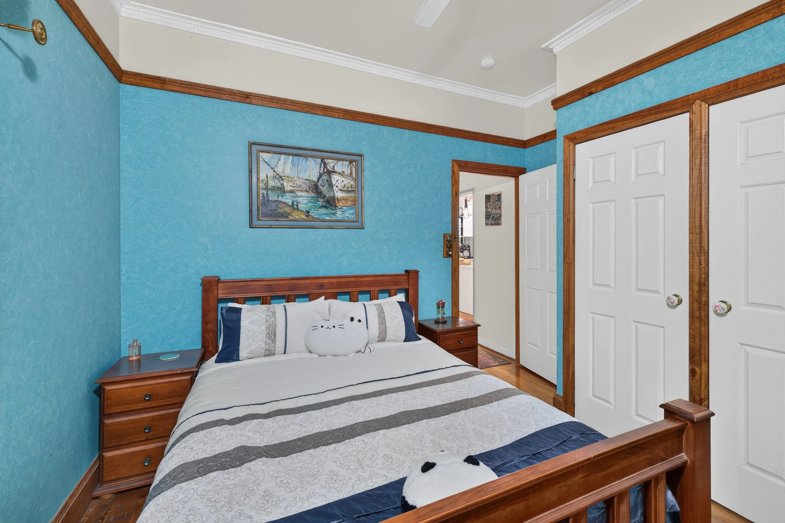 A bedroom with a blue textured wall, a wooden bed with a white bedspread and decorative pillows, a small nightstand with a vase of flowers and a lamp, a framed painting of boats on the wall, and a white closet door with wooden trim.