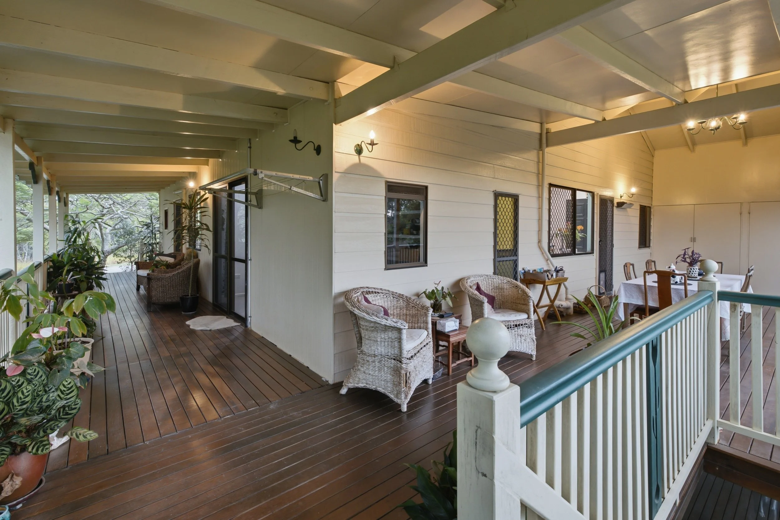 Covered porch with brown wooden flooring, white painted walls, multiple windows with black screens, and a dining table with chairs. There are wicker chairs and various potted plants along the edge of the porch. The ceiling has exposed beams and light