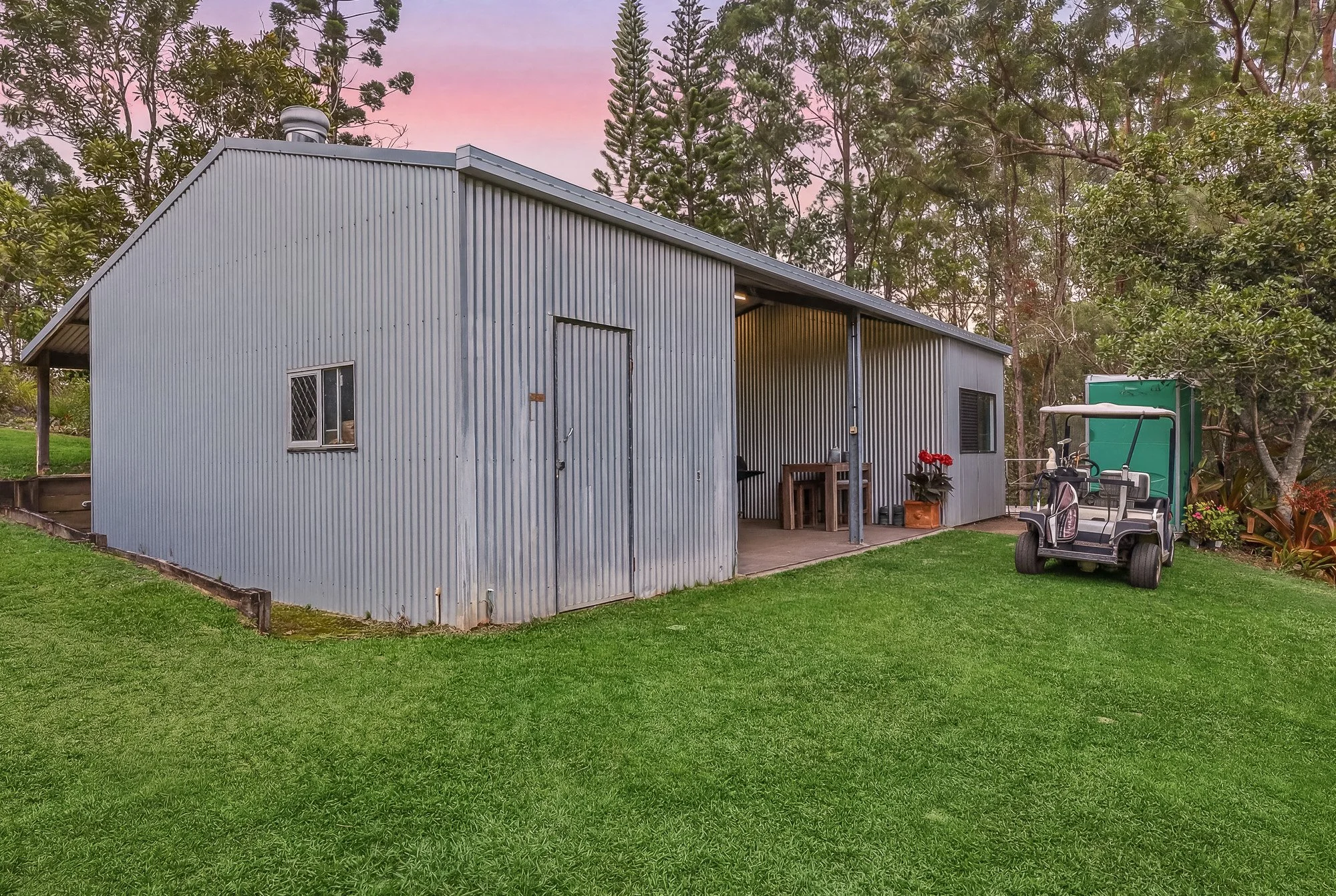 Photograph of a metal shed with a sloped roof, windows, and a small porch area, situated on a well-maintained grassy lawn with trees and bushes in the background during dusk. A golf cart is parked nearby.