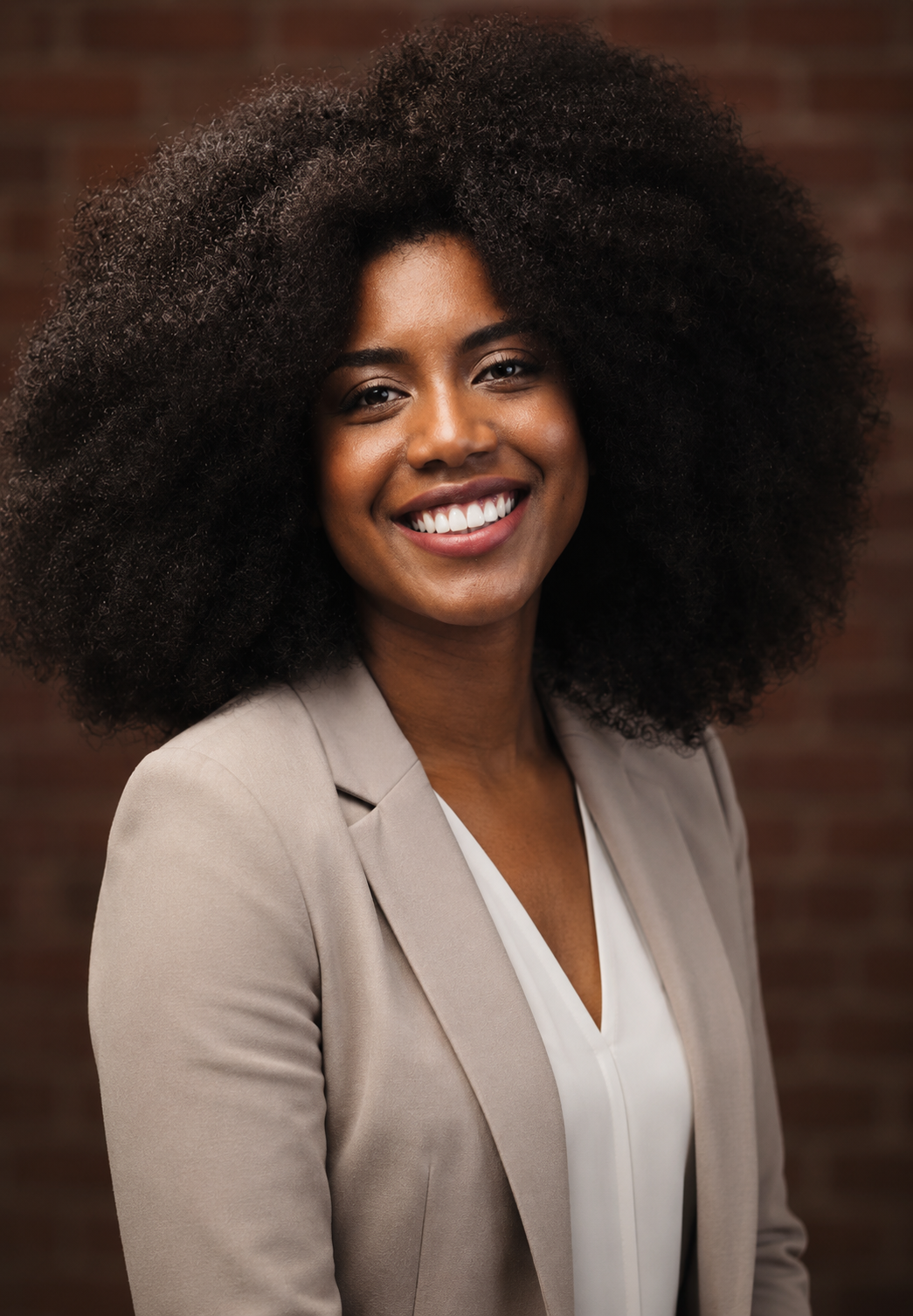 A woman with dark curly hair smiling, wearing a beige blazer and white blouse, standing against a brick wall background.