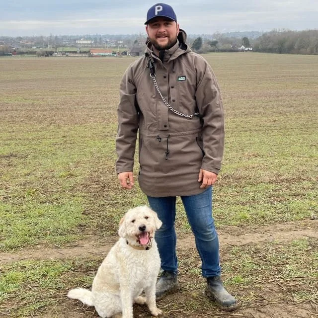 A man standing in a field with a small, fluffy white dog sitting beside him. The man is wearing a brown jacket, blue jeans, and a baseball cap.