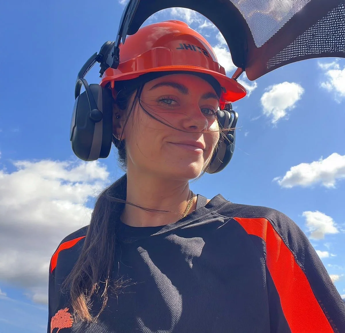 A woman wearing a safety helmet and noise-canceling headphones, smiling outdoors against a bright blue sky with clouds.