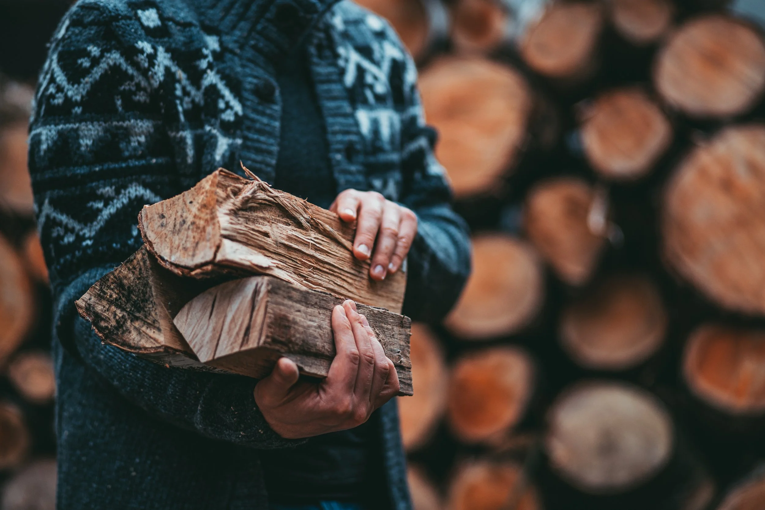 Person holding chopped firewood with a stack of logs in the background, wearing a knit sweater.