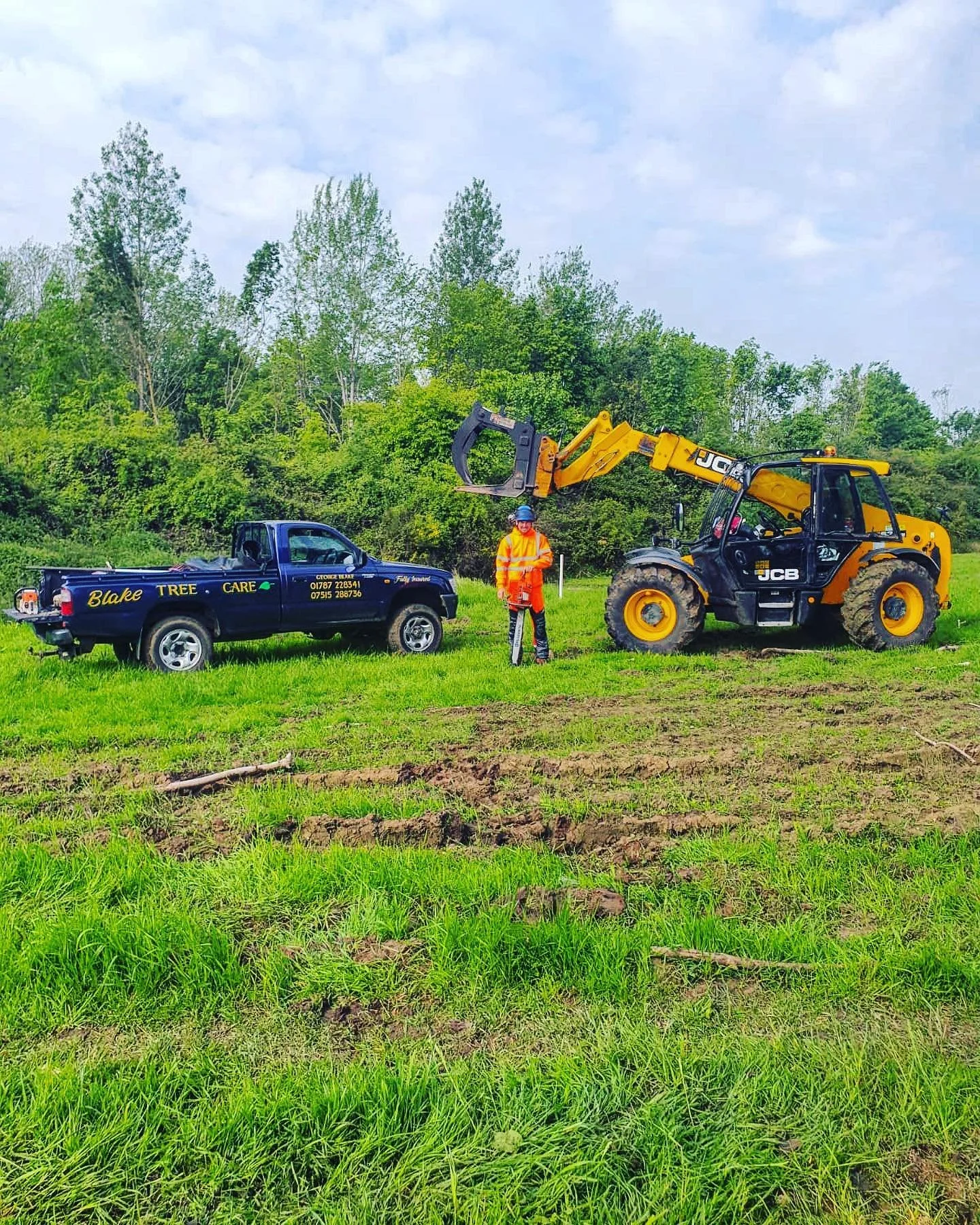 A worker in a bright orange safety jacket and blue helmet standing between a black and yellow JCB front loader and a dark blue truck in a grassy field with trees in the background.