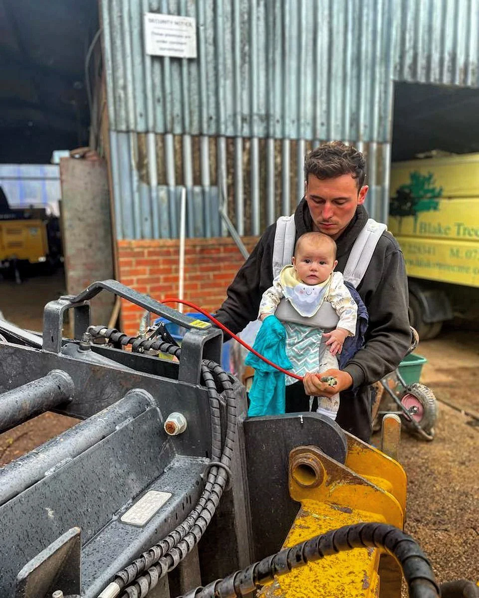 A man carries a baby in a front-facing baby carrier, inspecting construction machinery in a farm or industrial setting.