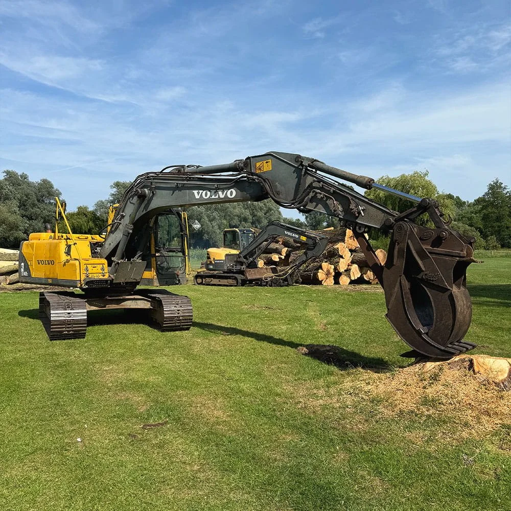 A large yellow and black Volvo excavator with a bucket attachment is on a grassy field, with a smaller excavator nearby and logs stacked in the background.