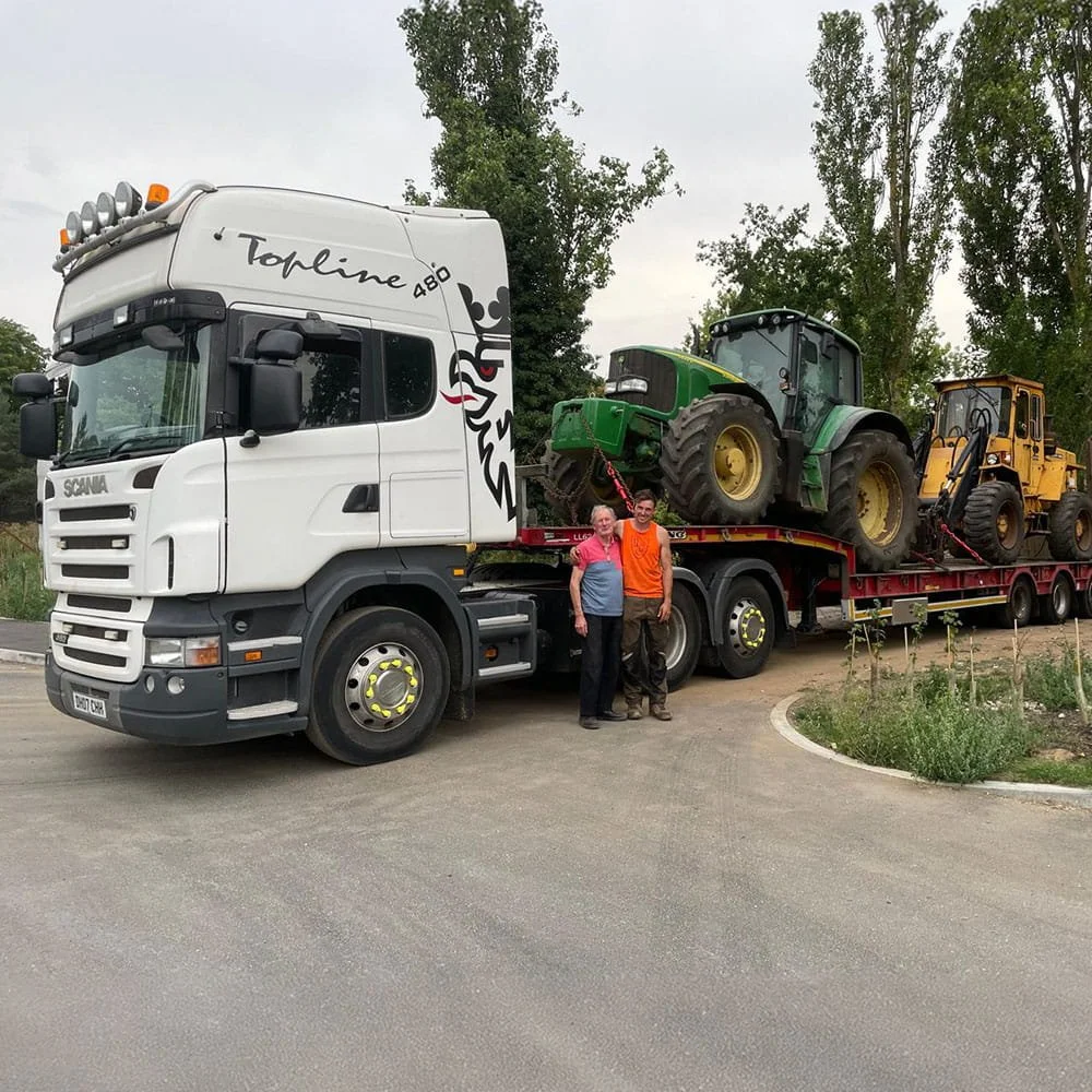 A white semi-truck with a trailer carrying a green tractor and a yellow construction vehicle parked on a curved road with trees in the background. Two people are standing in front of the truck, smiling for the photo.