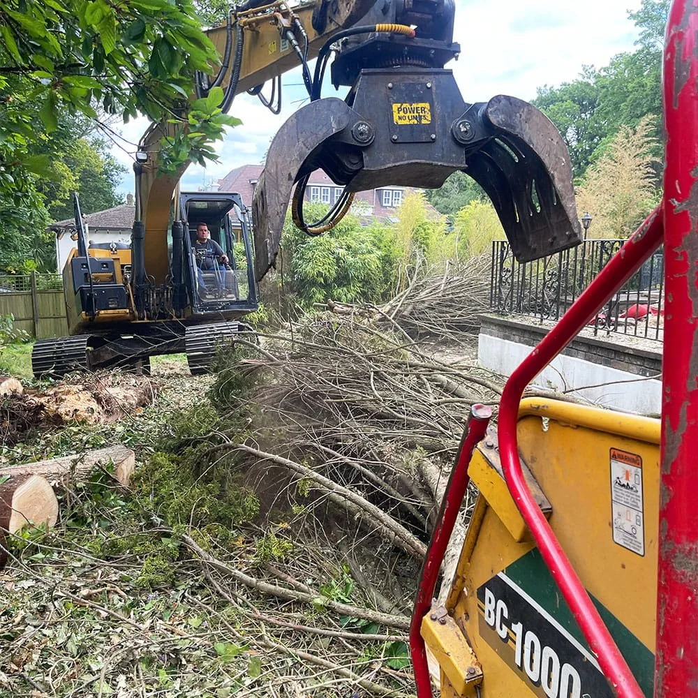 A construction worker operating a large excavator with a claw attachment, clearing fallen tree branches and debris from a yard near a house.
