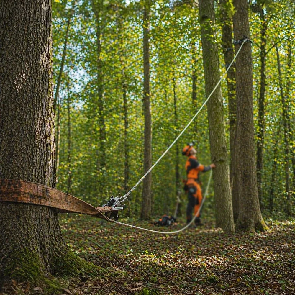 A person in orange safety gear and helmet working on tree climbing equipment attached to a tree in a dense forest.