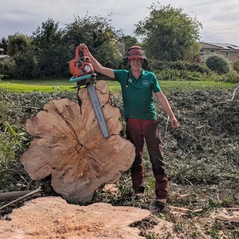 A woman standing outdoors next to a large fallen tree trunk, holding a chainsaw, wearing a helmet with ear and face protection, a green shirt, and pants.