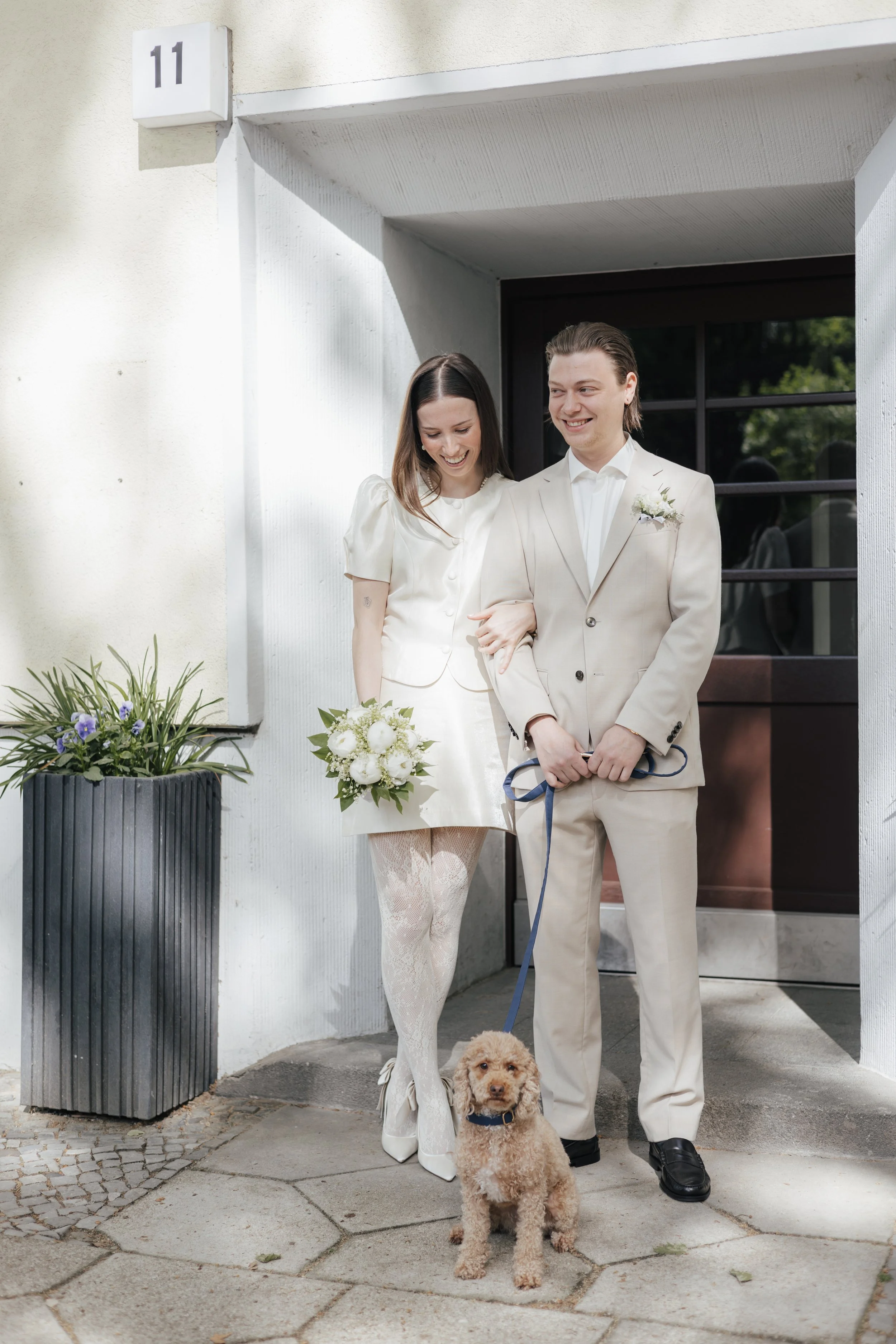 Ein Paar in Hochzeitskleidung steht vor dem Eingang eines Hauses, hält Händchen und lächelt. Ein kleiner brauner Hund sitzt vor ihnen auf dem Boden. Es sind Blumen und Pflanzen neben der Tür zu sehen, die Hausnummer ist 11.