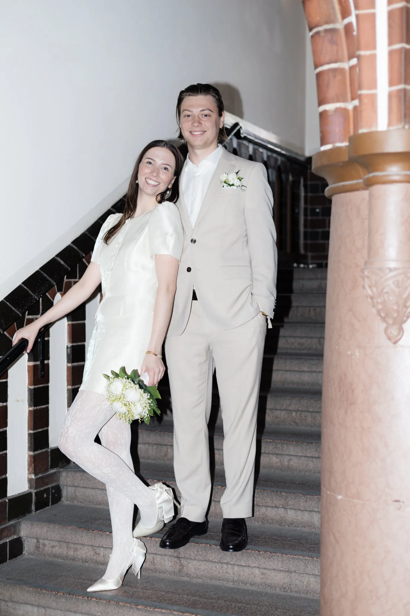 Ein Brautpaar steht auf einer Treppe in einem Haus, lächelnd in die Kamera. Die Frau trägt ein weißes Hochzeitskleid mit Spitze und weiße High Heels, hält einen Brautstrauß. Der Mann trägt einen hellbeigen Anzug mit Hemd und hat eine kleine weiße Blu