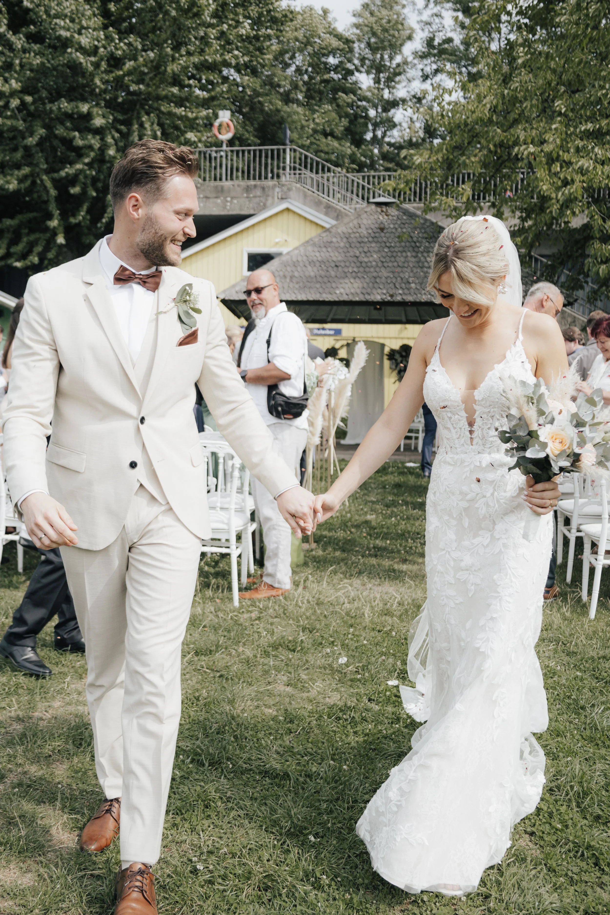 Ein Brautpaar tanzt auf einer grünen Wiese bei einer Outdoor-Hochzeit. Der Bräutigam trägt einen weißen Anzug mit braunem Hemd und Schleife, die Braut trägt ein weißes Hochzeitskleid und hält einen Blumenstrauß. Im Hintergrund sind weitere Gäste und 