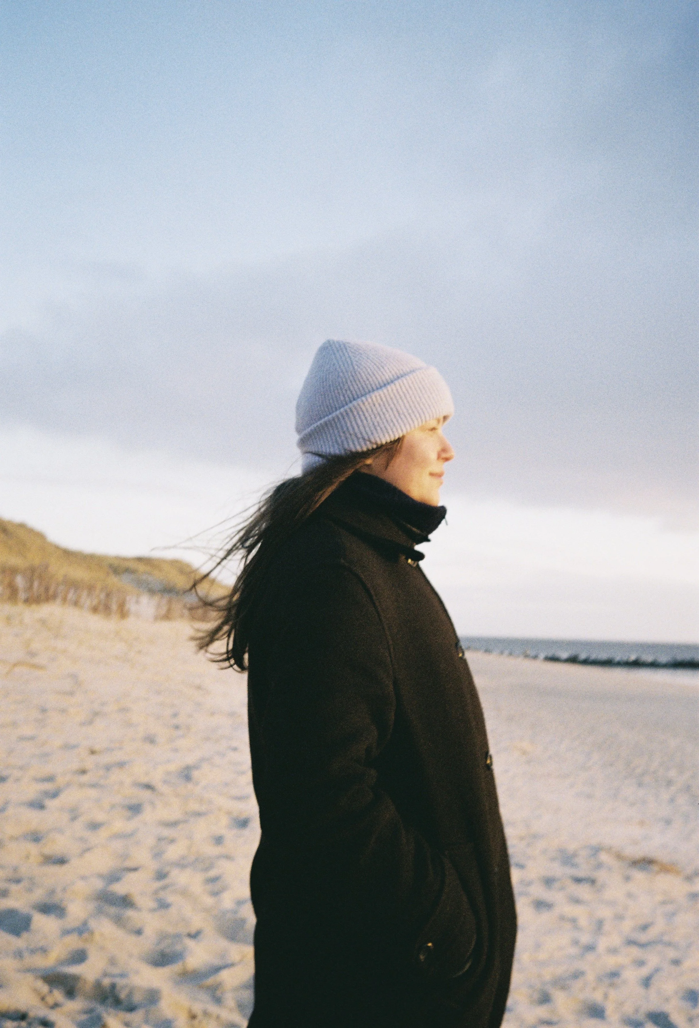 Eine Frau steht am Strand bei Sonnenuntergang, trägt eine graue Mütze und eine schwarze Jacke, blickt nach rechts.