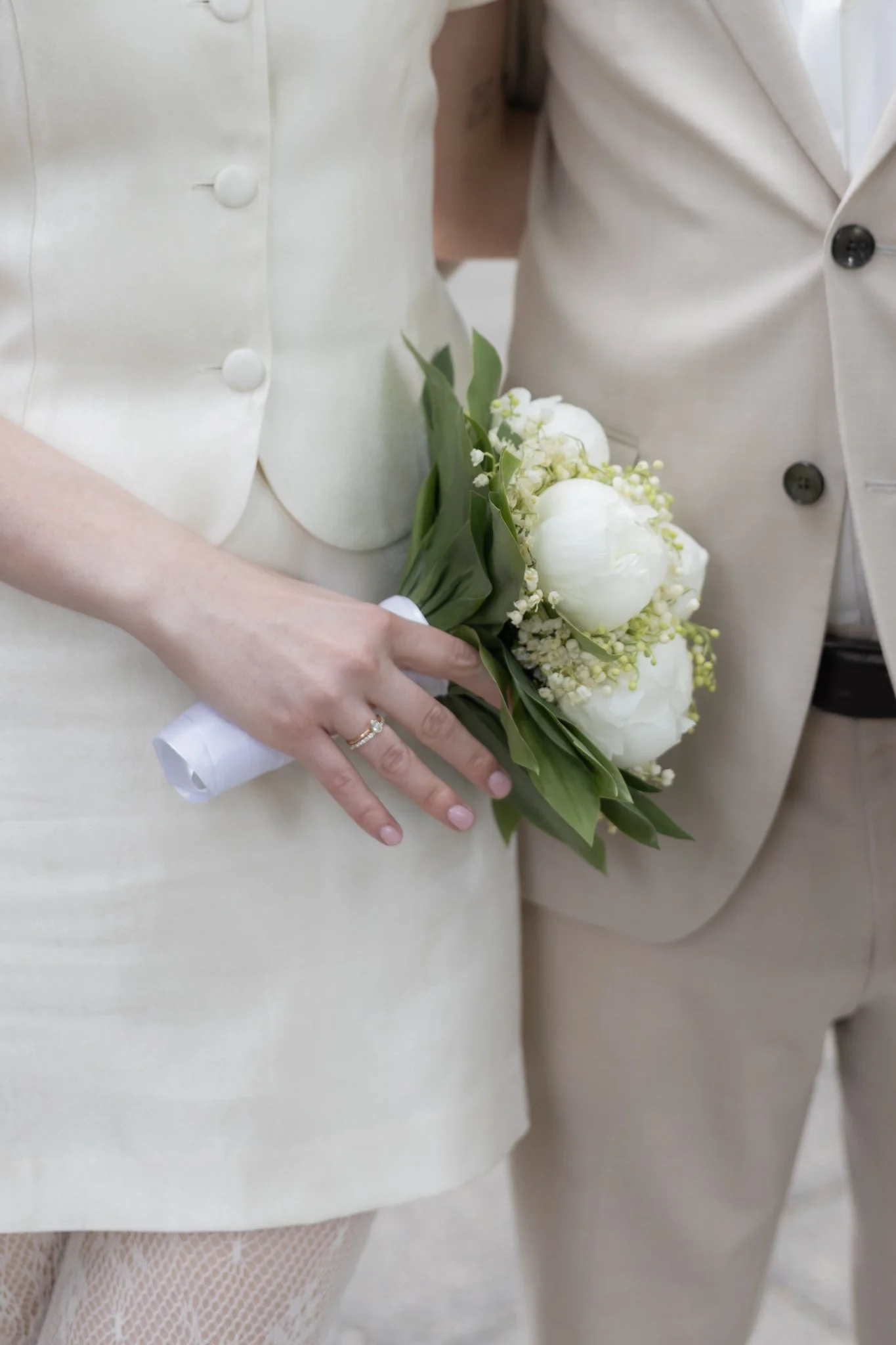 Nahaufnahme von einer Frau, die ein kleines Bouquet aus weißen Blumen hält, während sie ein weißes Kleid trägt, vermutlich bei einer Hochzeit.
