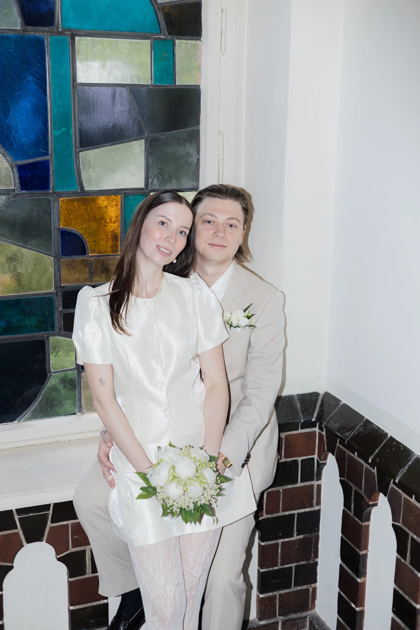 Ein Paar bei einer Hochzeit, im Hintergrund ein buntes Glasfenster, sie halten einen Brautstrauß, beide tragen helle Kleidung und posieren vor einer weißen Wand und einer dunklen Samtbrüstung.