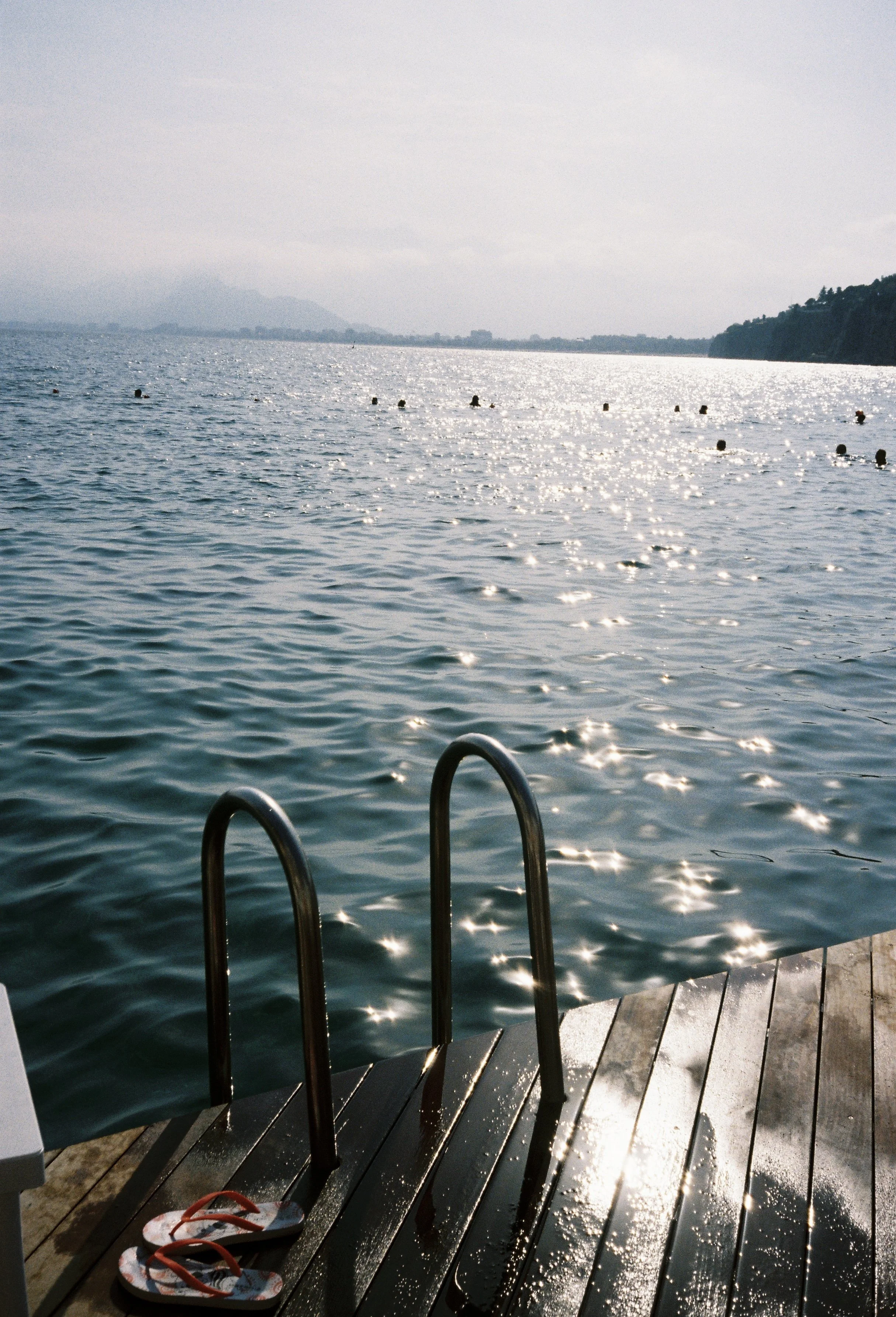 Ein Steg am Wasser mit einer Leiter, um ins Wasser zu gelangen. Im Wasser sind Bojen, im Hintergrund Berge und eine bewölkte Himmel. Auf dem Steg liegen Flip-Flops.