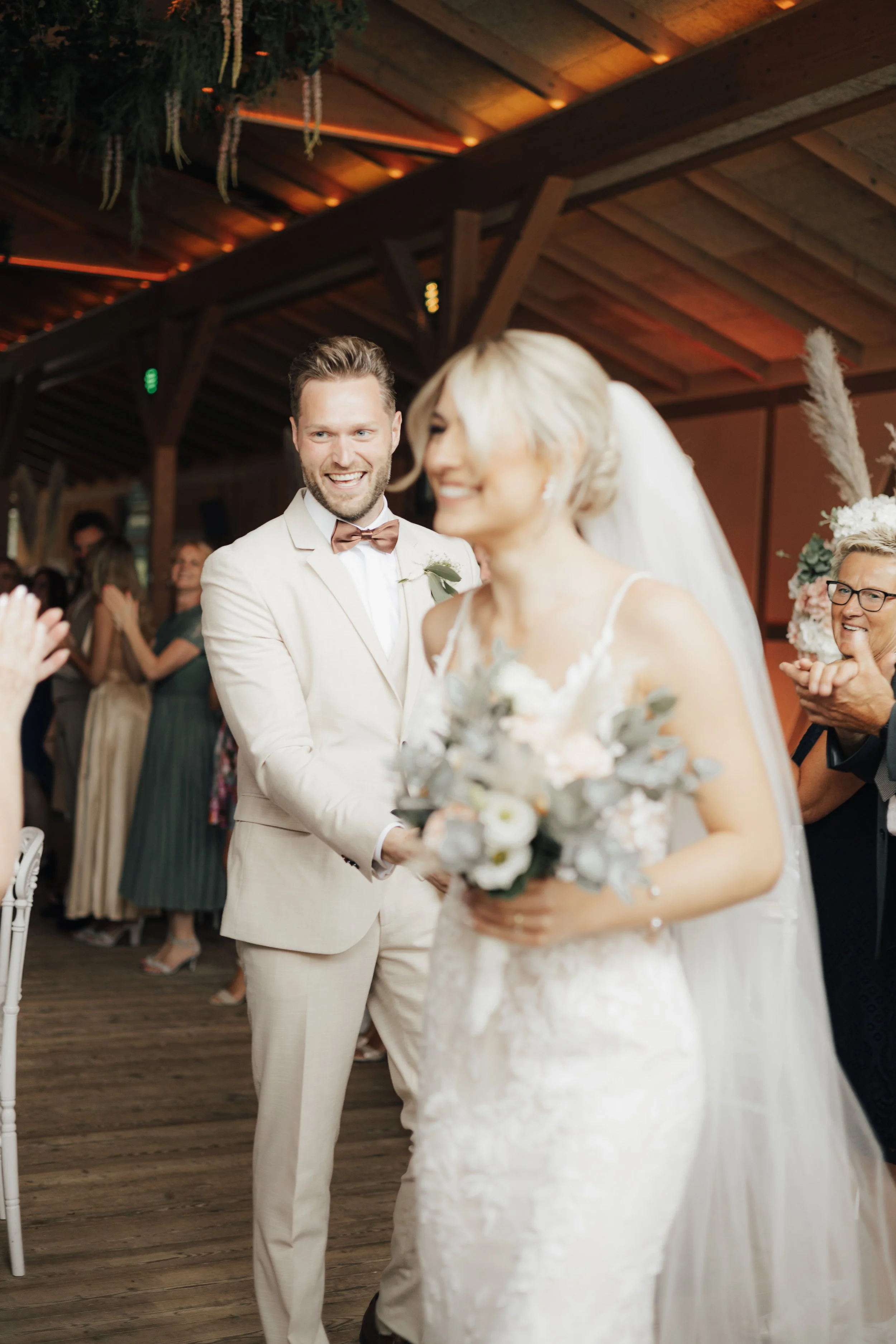 Ein Brautpaar bei ihrer Hochzeit, der Bräutigam lacht, während die Braut mit einem Blumenstrauß vorbeigeht, im Hintergrund singen Gäste in einer rustikalen Holzhütte.