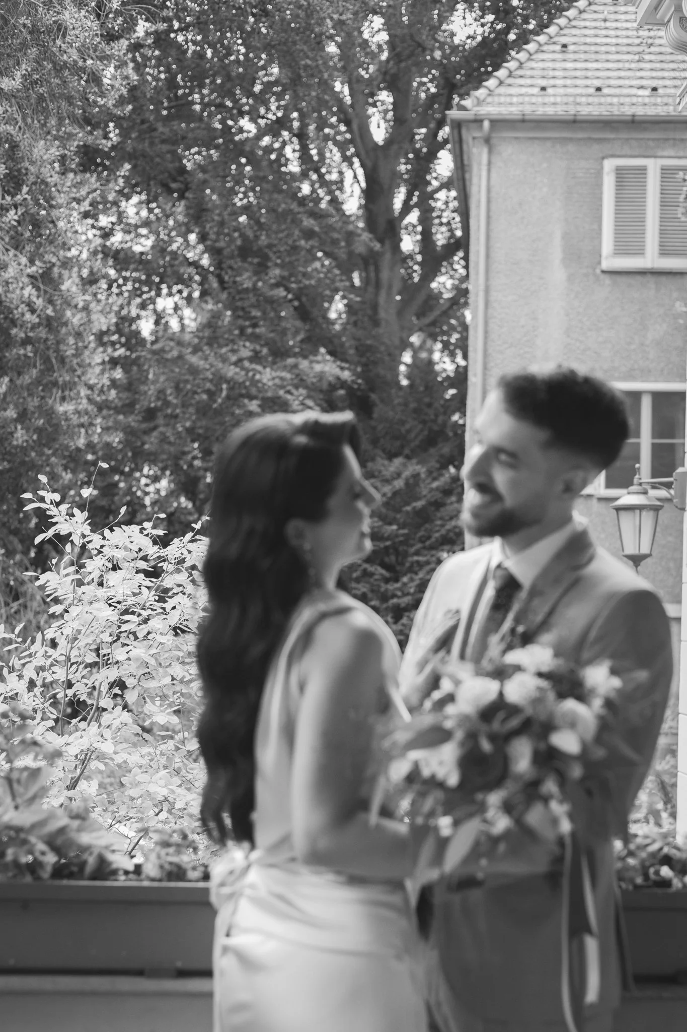Ein Mann und eine Frau sehen sich lächelnd an, während sie sich in einem Garten mit Bäumen und einem Haus im Hintergrund befinden. Der Mann hält einen Blumenstrauß.