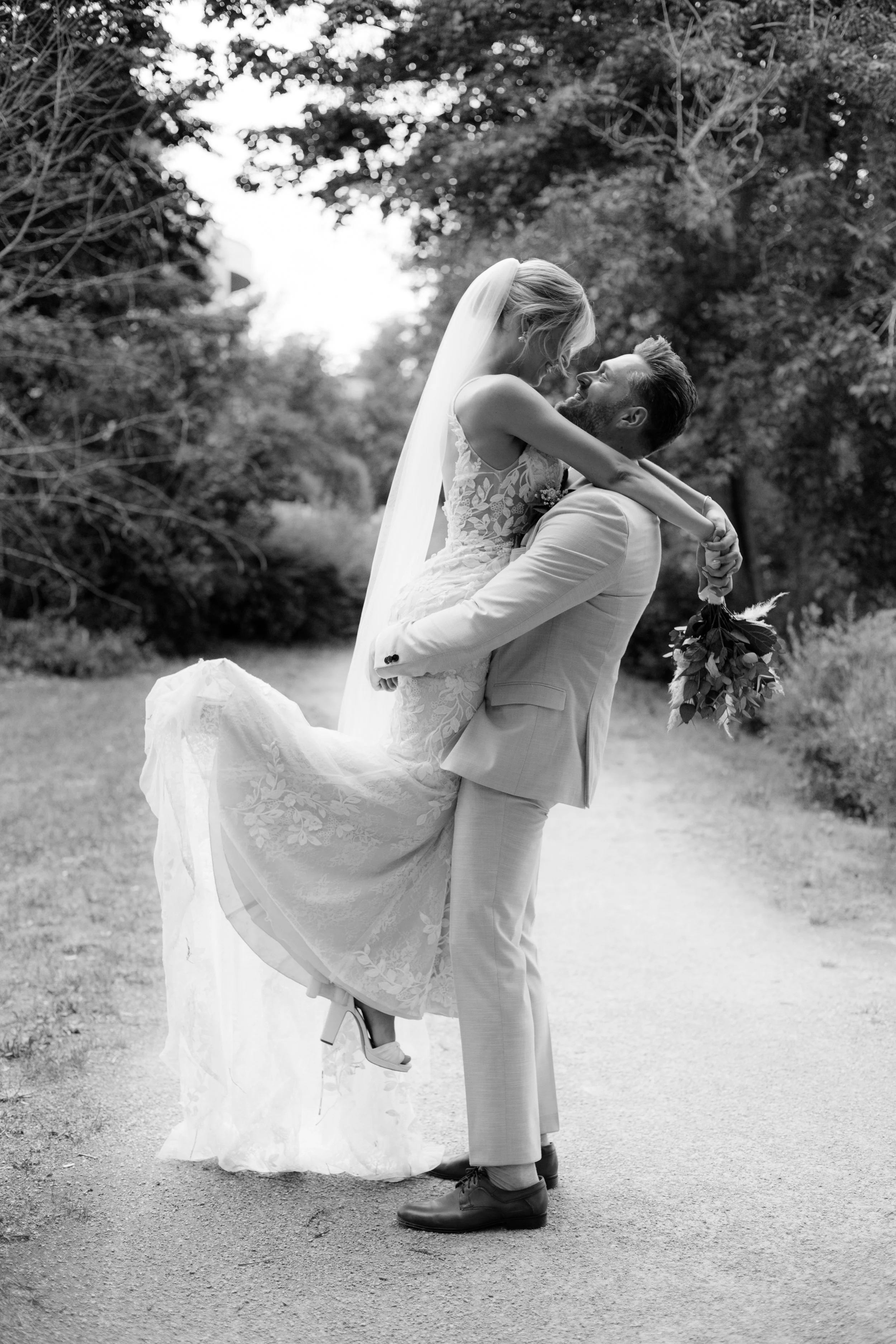 Ein Brautpaar bei einer Hochzeit im Freien, der Bräutigam hebt die Braut hoch, sie trägt ein Hochzeitskleid und einen Schleier, er trägt einen Anzug, sie hält einen Blumenstrauß, im Hintergrund Bäume und natürlicher Hintergrund.