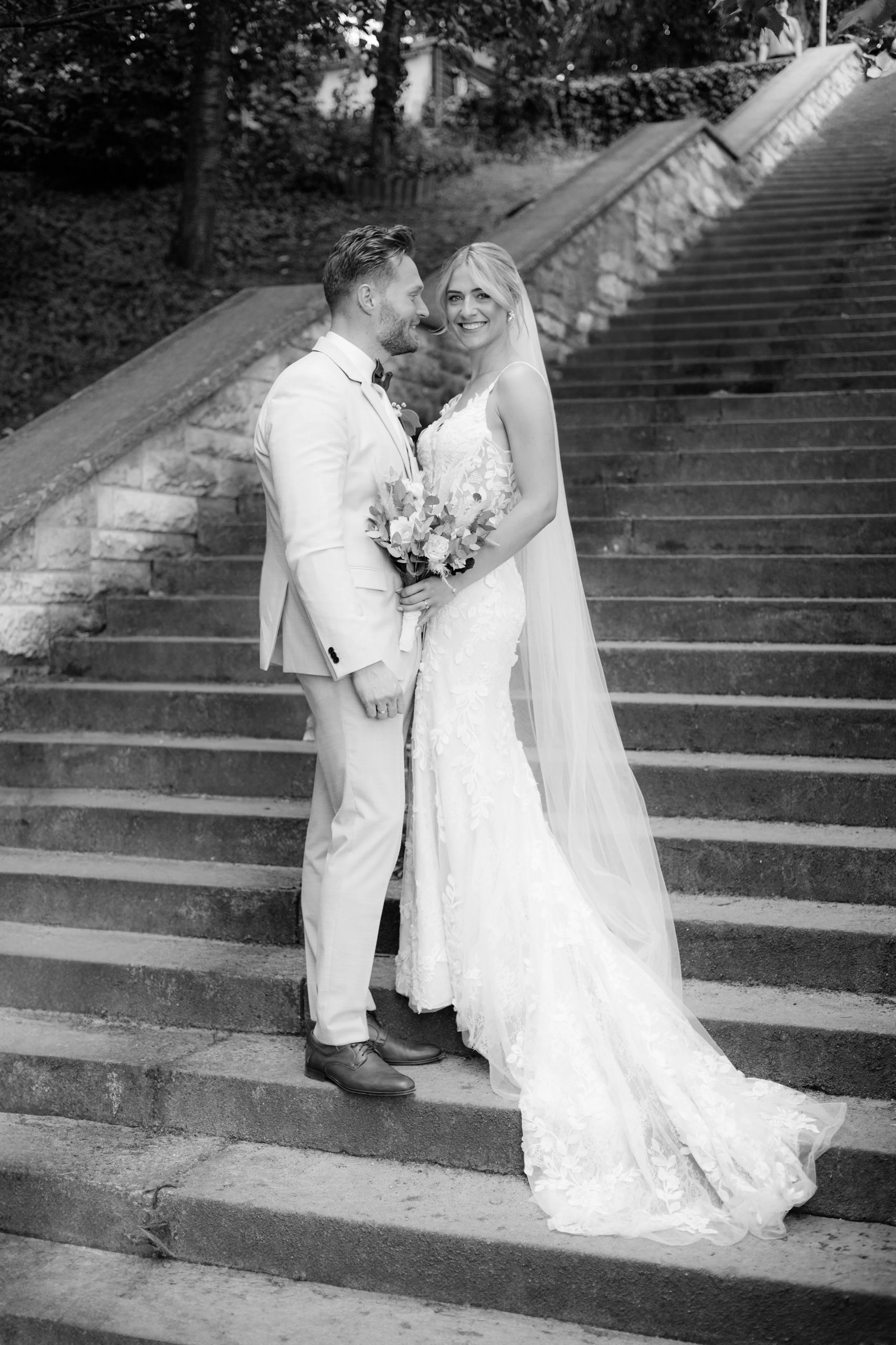 Ein Schwarz-Weiß-Foto eines Brautpaares auf einer steinernen Treppe im Freien, das Braut trägt ein langes, Spitze Brautkleid mit Schleier und hält einen Blumenstrauß, der Bräutigam trägt einen hellen Anzug mit Fliege.