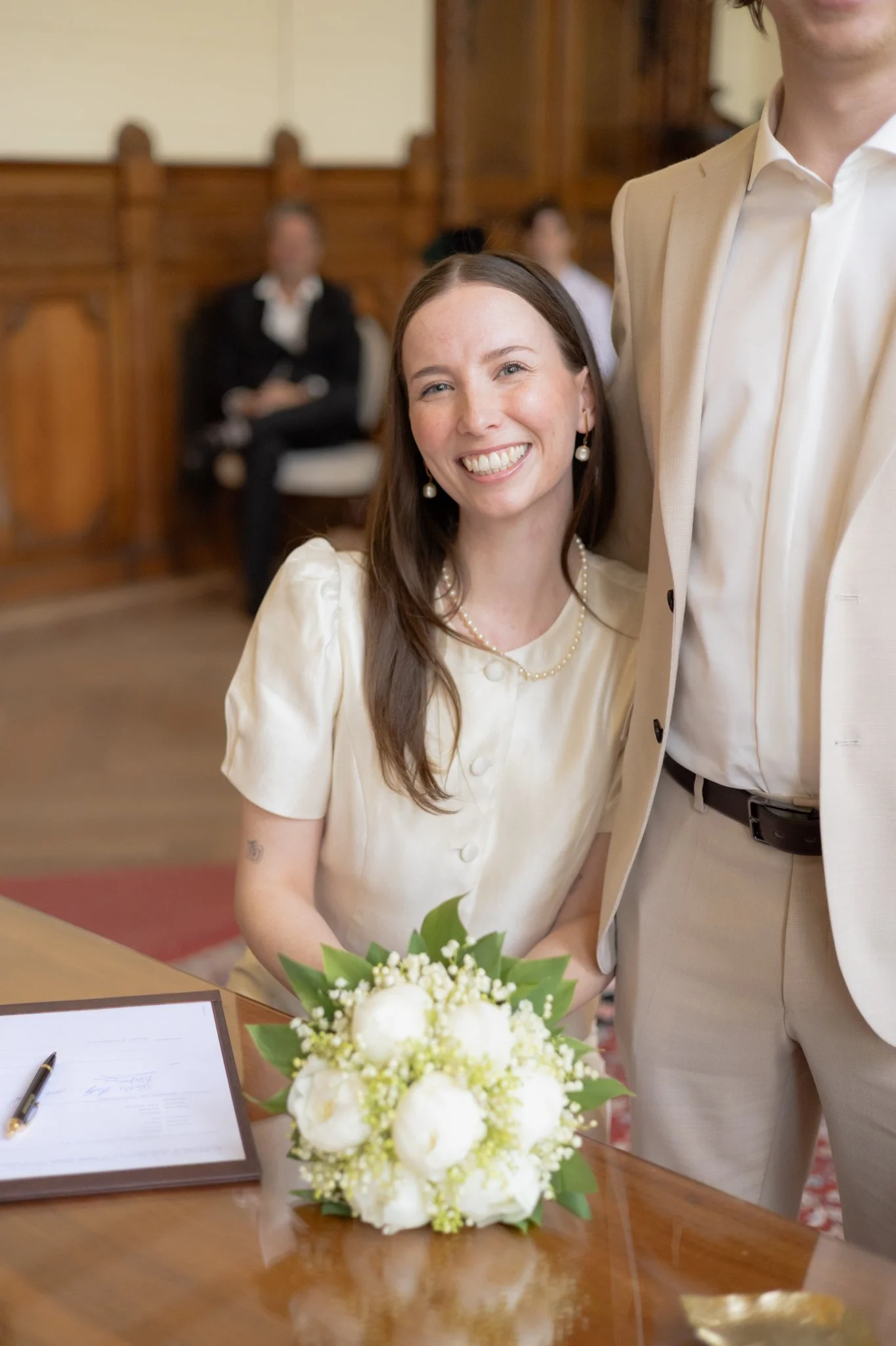 Eine Frau mit braunen Haaren, die Freude zeigt, sitzt an einem Tisch bei einer Zeremonie, wahrscheinlich einer Hochzeit, mit einem Blumenstrauß vor sich. Im Hintergrund sitzen Menschen in einem Raum mit holzvertäfelten Wänden.