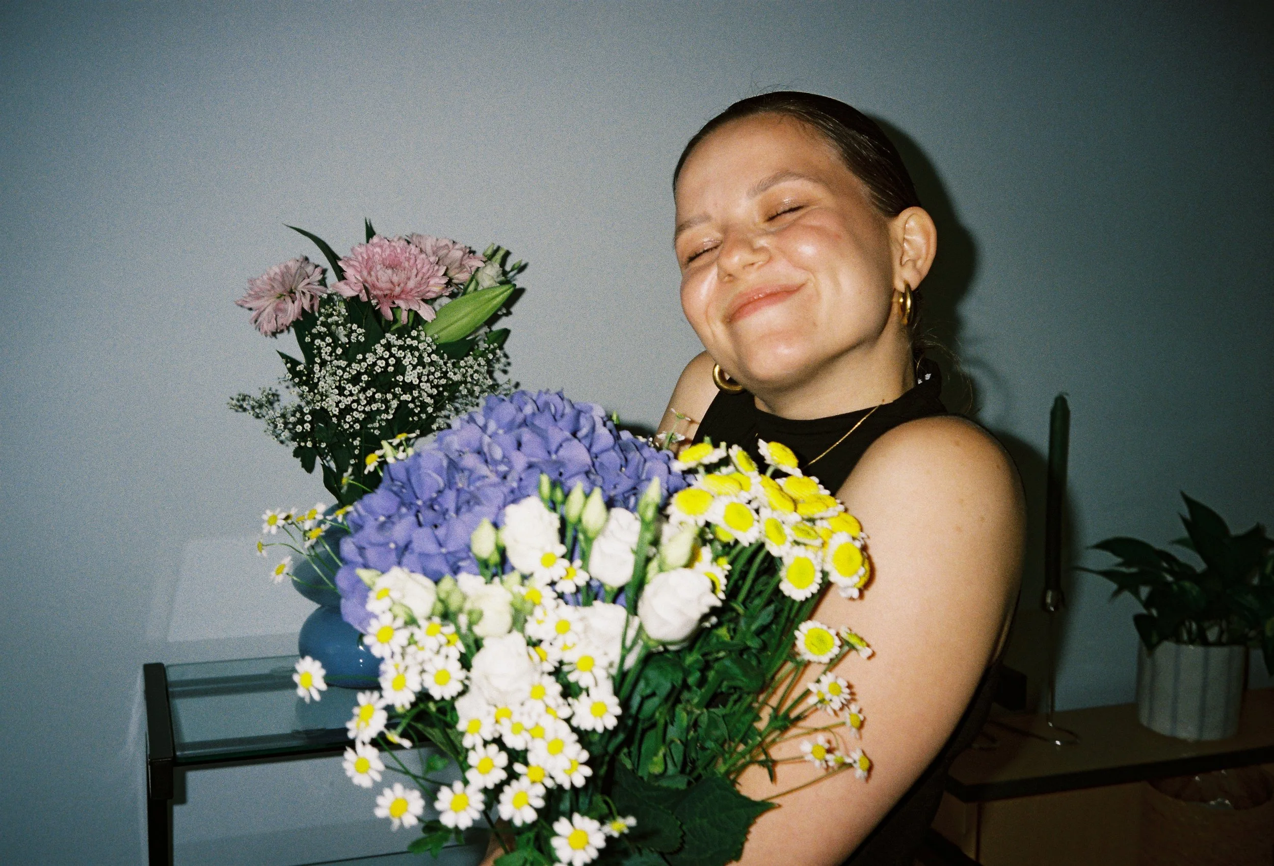 Eine lachende Frau mit dunklen, glatten Haaren, schulterlang, hält einen großen Blumenstrauß mit pinken, lila, weißen und gelben Blumen, vor einem grauen Hintergrund.