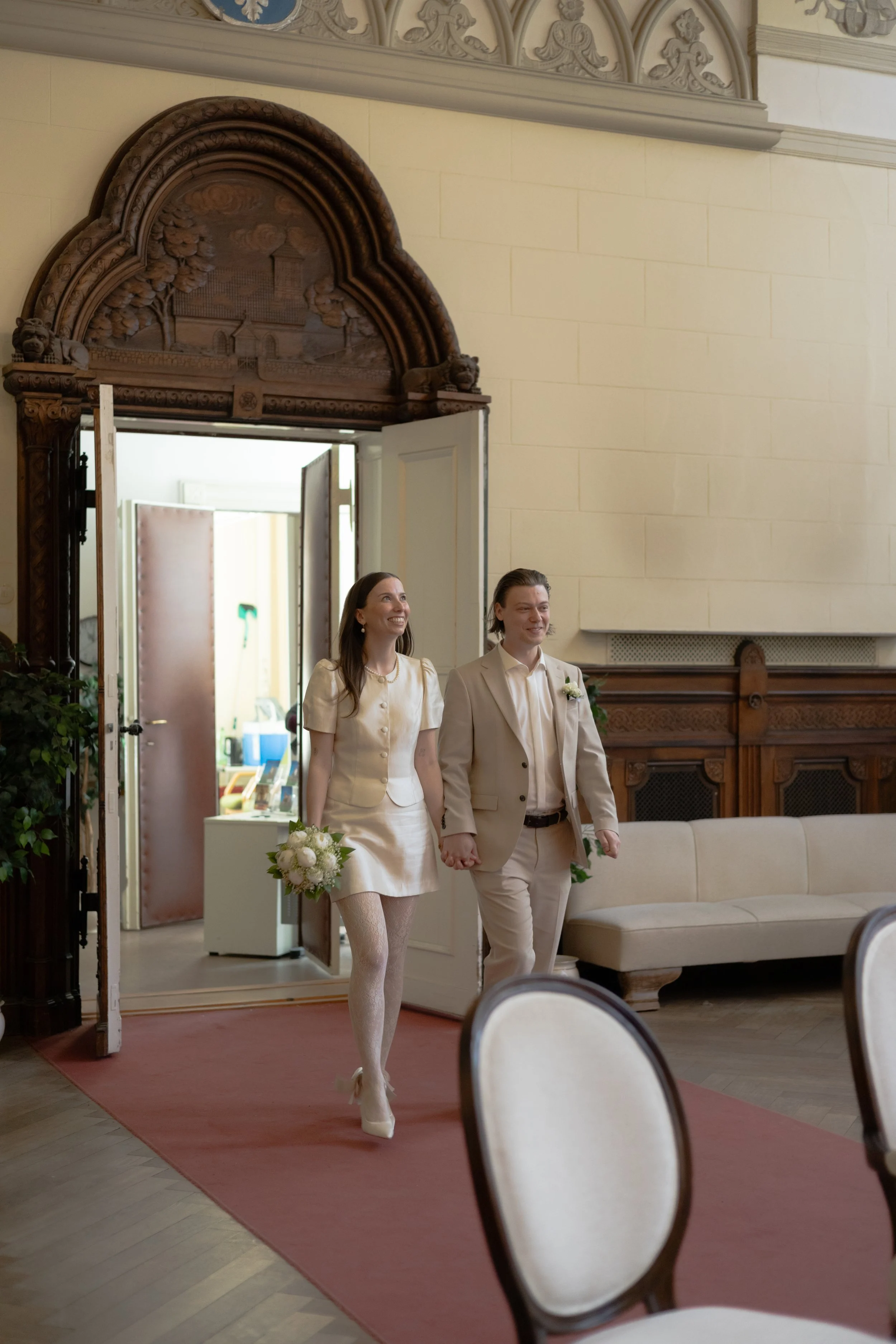 Ein frisch verheiratetes Paar, das die Hand hält, in einem eleganten Saal bei einer Hochzeit. Die Frau trägt ein weißes Kleid mit Strumpfhosen und Pumps, die Männer trägt einen hellen Anzug mit Blumen im Revers.