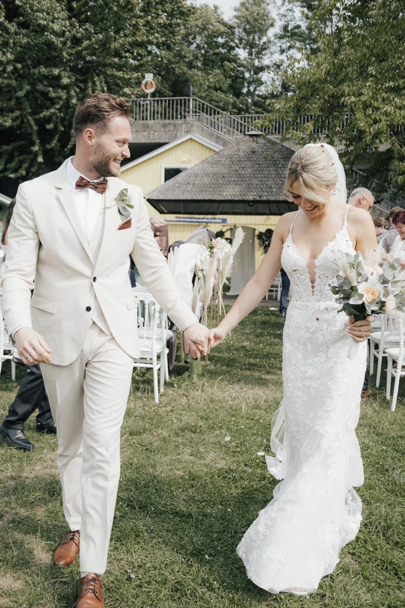 Ein Brautpaar hält Händchen und lacht bei einer Hochzeitsfeier im Freien. Der Bräutigam trägt einen weißen Anzug mit braunem Bowtie und braunen Schuhen, die Braut trägt ein weißes, florales Hochzeitskleid mit Schleier und hält einen Blumenstrauß. Im 