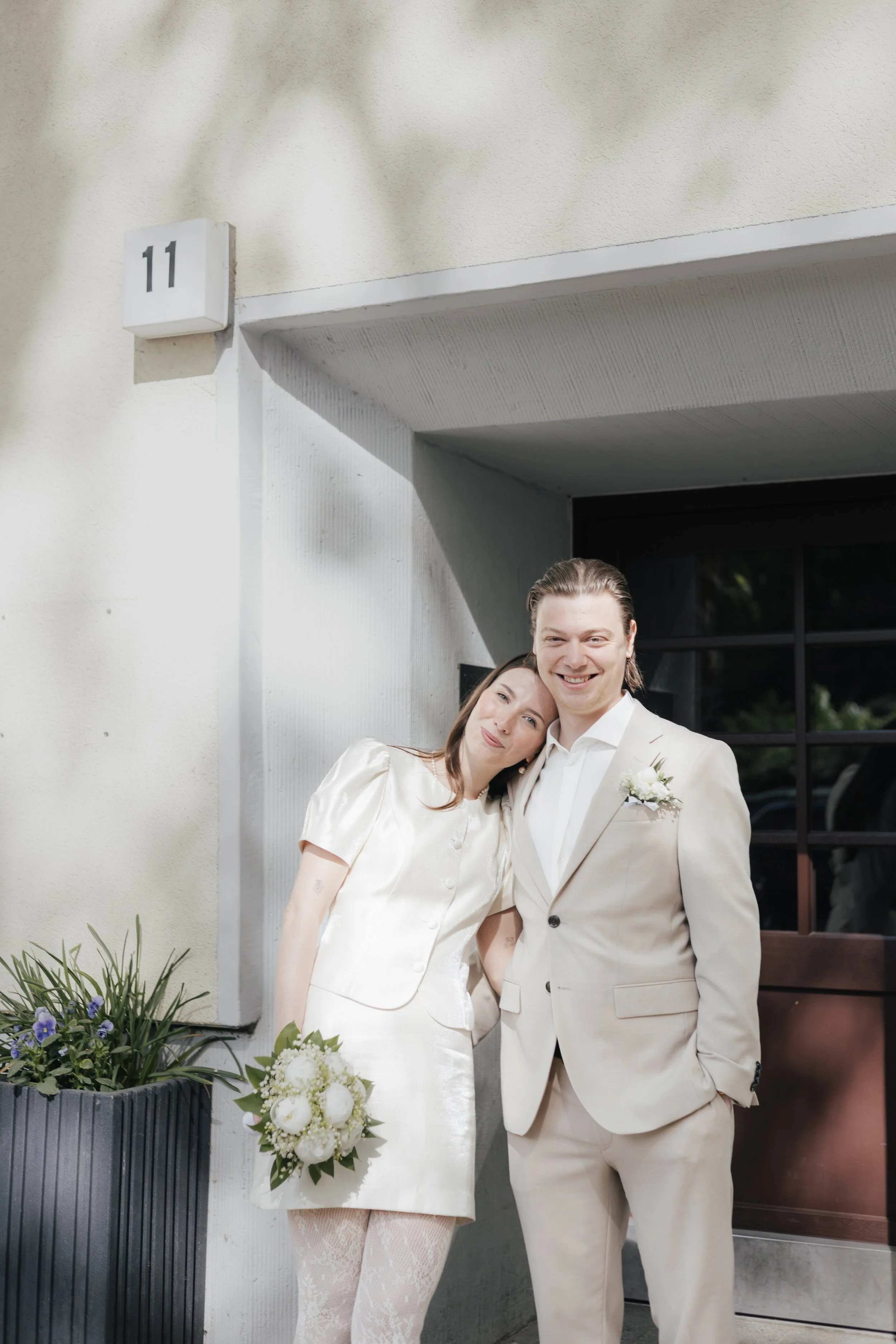 Ein glückliches Brautpaar steht vor einem weißen Haus, umgeben von Pflanzen und Blumen, an einem sonnigen Tag, bei ihrer Hochzeit.