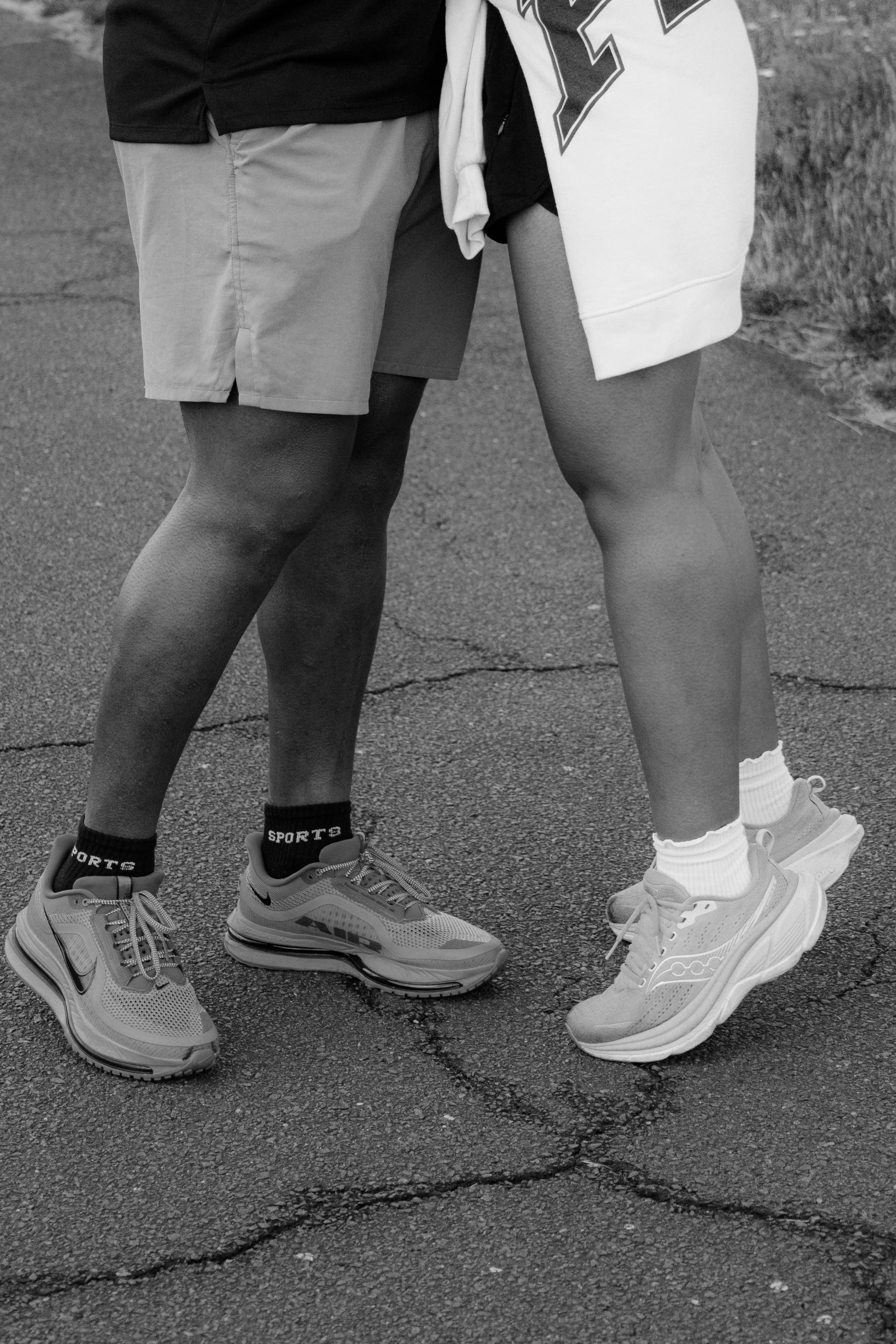 Vor einem dunklen Asphaltboden stehen zwei Menschen mit unterschiedlichen Hautfarben, nur ihre Beine und Schuhe sind sichtbar. Einer trägt helle Sneakers, schwarze Socken mit dem Schriftzug "SPORTS", Shorts, ein langärmliches schwarzes Oberteil und e