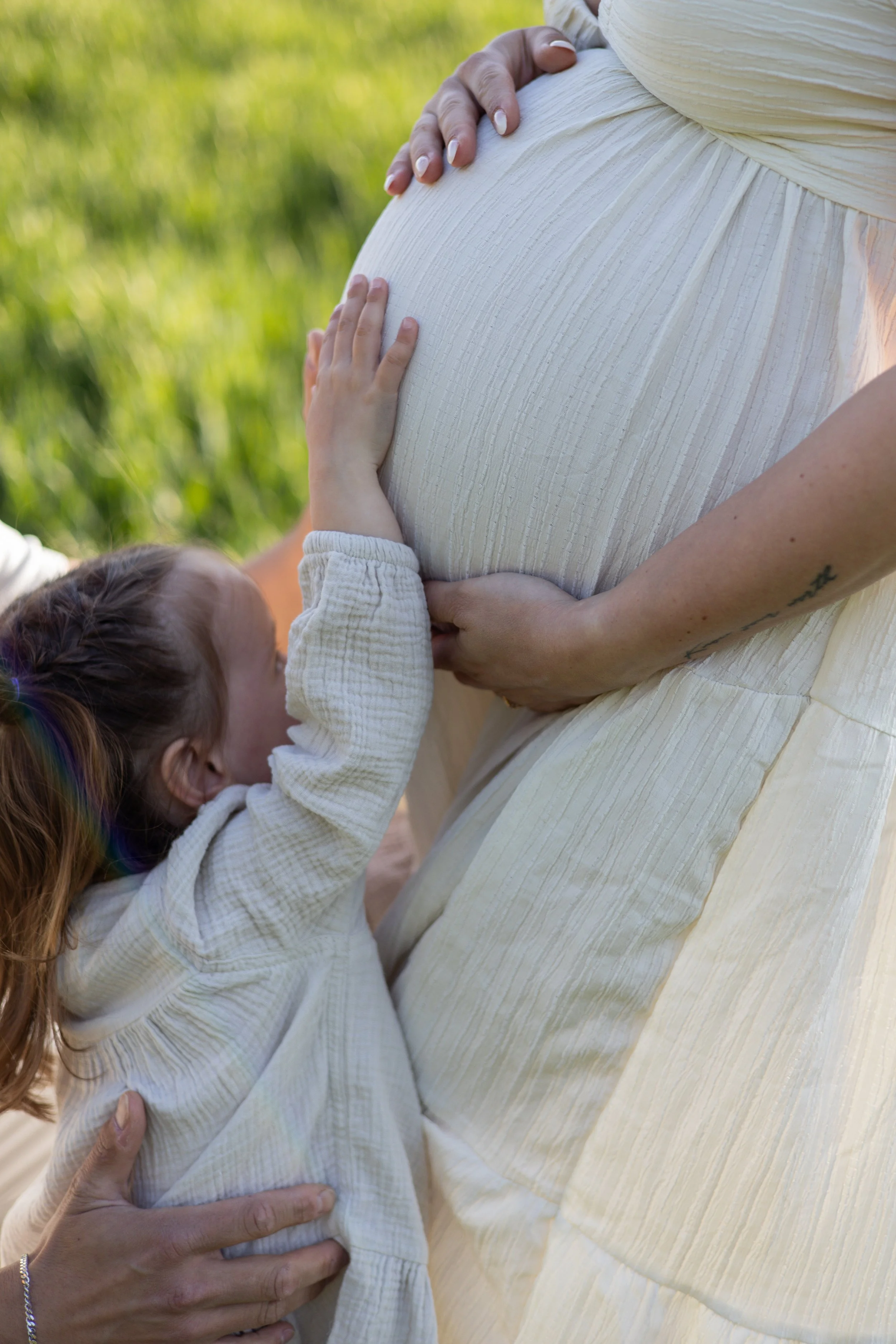 Eine schwangere Frau in einem weißen Kleid wird von einem Kind an ihrem Bauch gestreichelt, im Hintergrund ist grünes Gras sichtbar.
