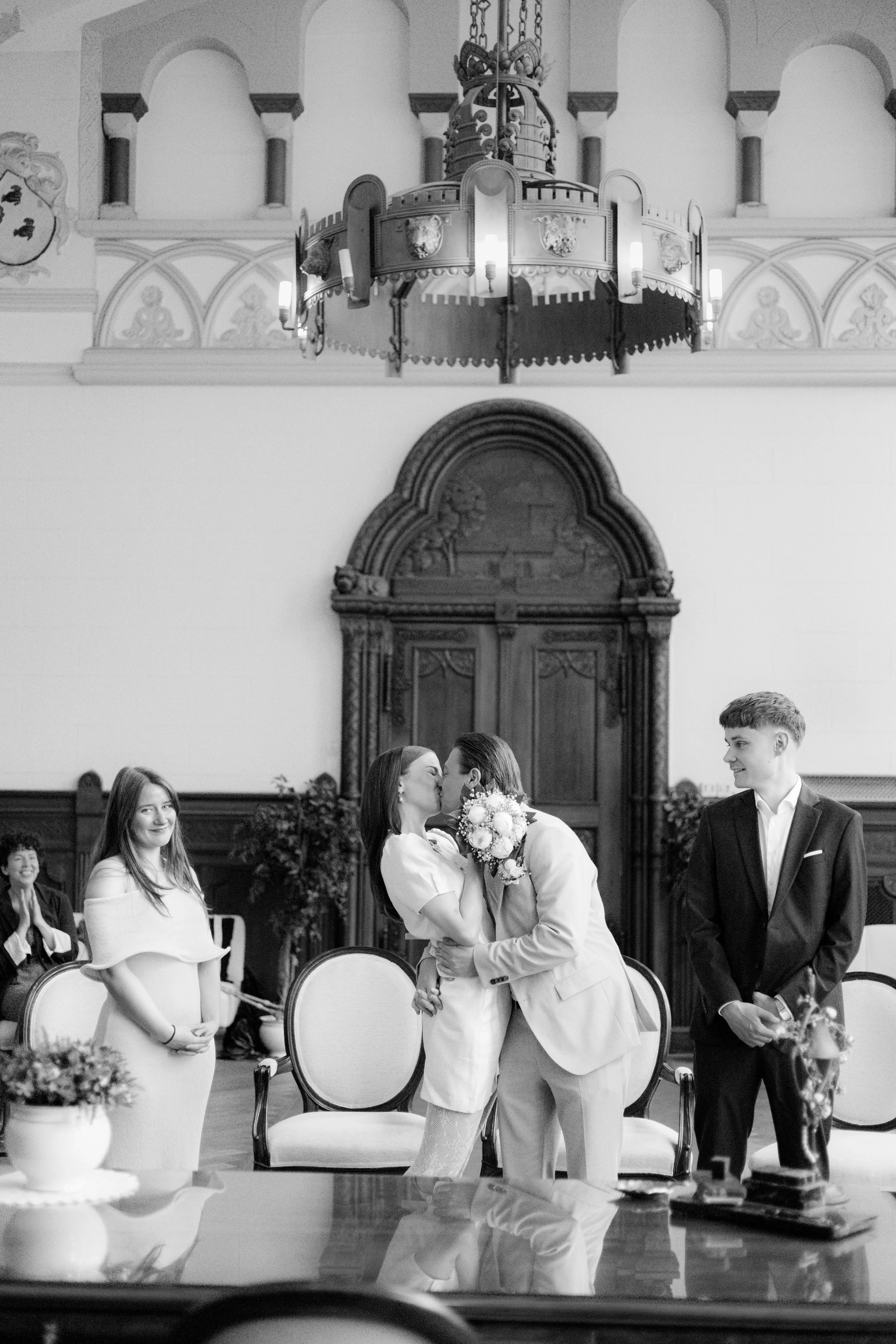 Ein Hochzeitspaar im festlichen Rahmen, umgeben von Gästen, im Innenraum eines prunkvollen Saals mit sehr hohen Decken und sehr dekorativen Wänden, die zur Zeremonie zeigen.