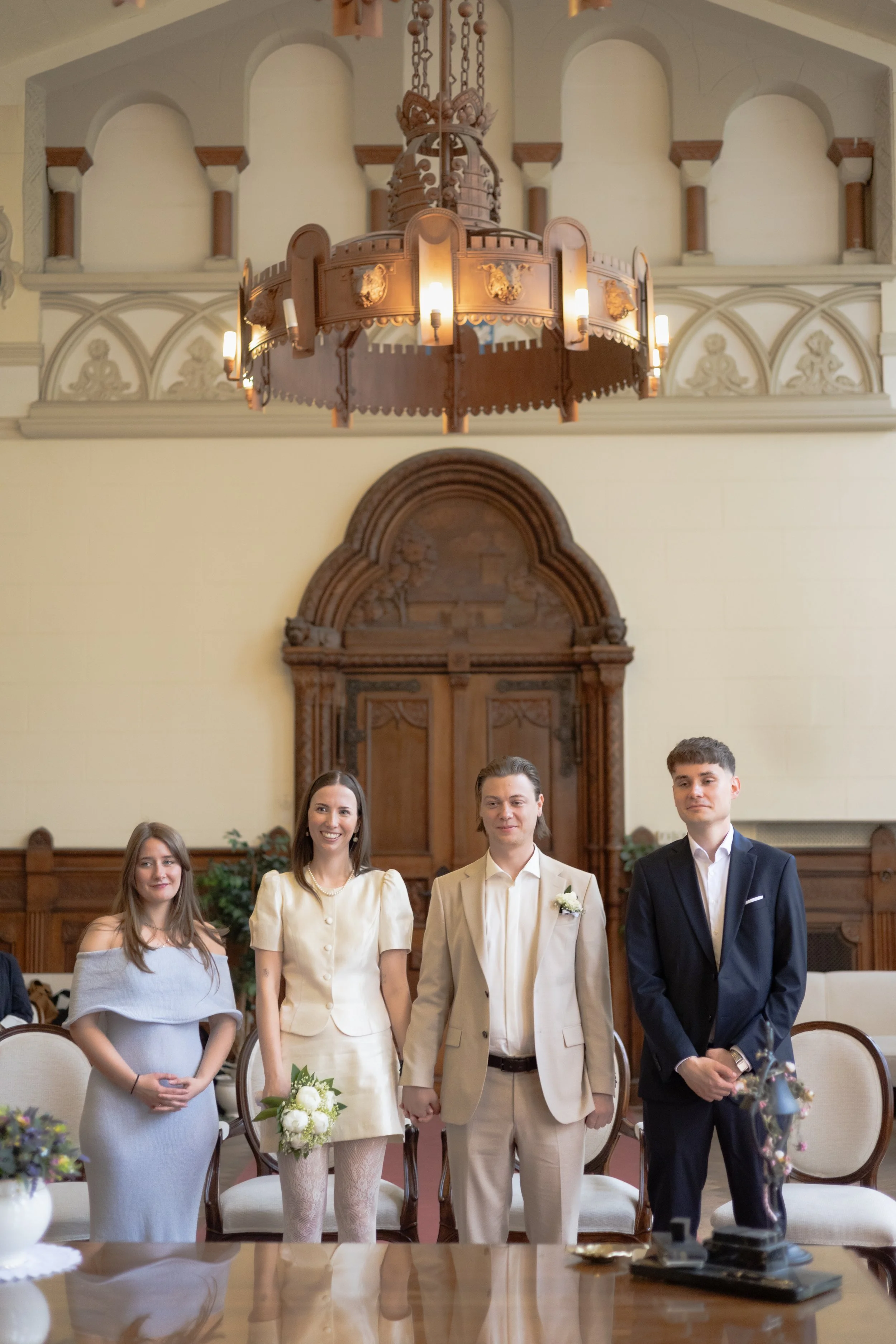 Fünf junge Erwachsene bei einer Hochzeitsveranstaltung in einem eleganten Raum mit Holzdekor. Das Brautpaar steht in der Mitte, die Frauen tragen elegante Kleider, die Männer Anzüge. Es sieht nach einer feierlichen Zeremonie aus.