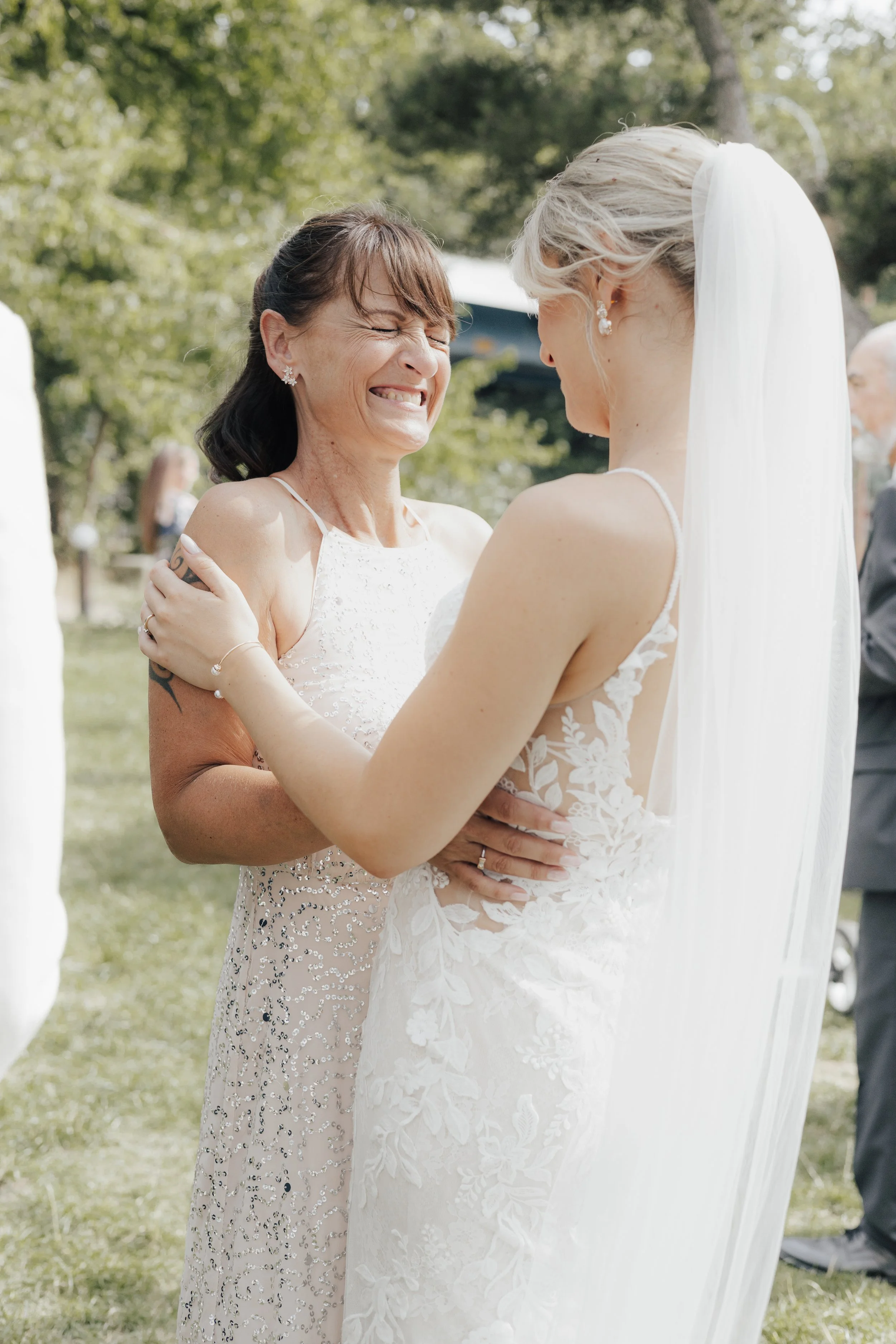 Eine emotionale Szene bei einer Hochzeit, bei der eine ältere Frau und eine junge Braut sich herzlich umarmen. Die Frau trägt ein hellrosafarbenes, perlenbesetztes Kleid und hat dunkle Haare, während die Braut einen weißen Hochzeitsanzug mit Spitzenm