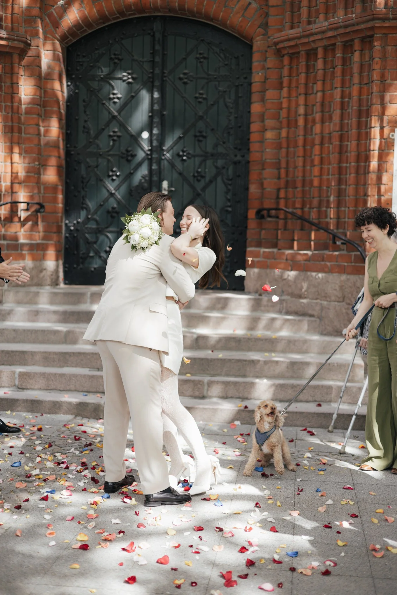 Ein Paar in Hochzeitskleidung umarmt sich vor einer roten Backsteinfassade, während Gäste Blumenblätter werfen. Ein kleines Hund trägt einen Pullover, und eine Frau mit Krücken lächelt.