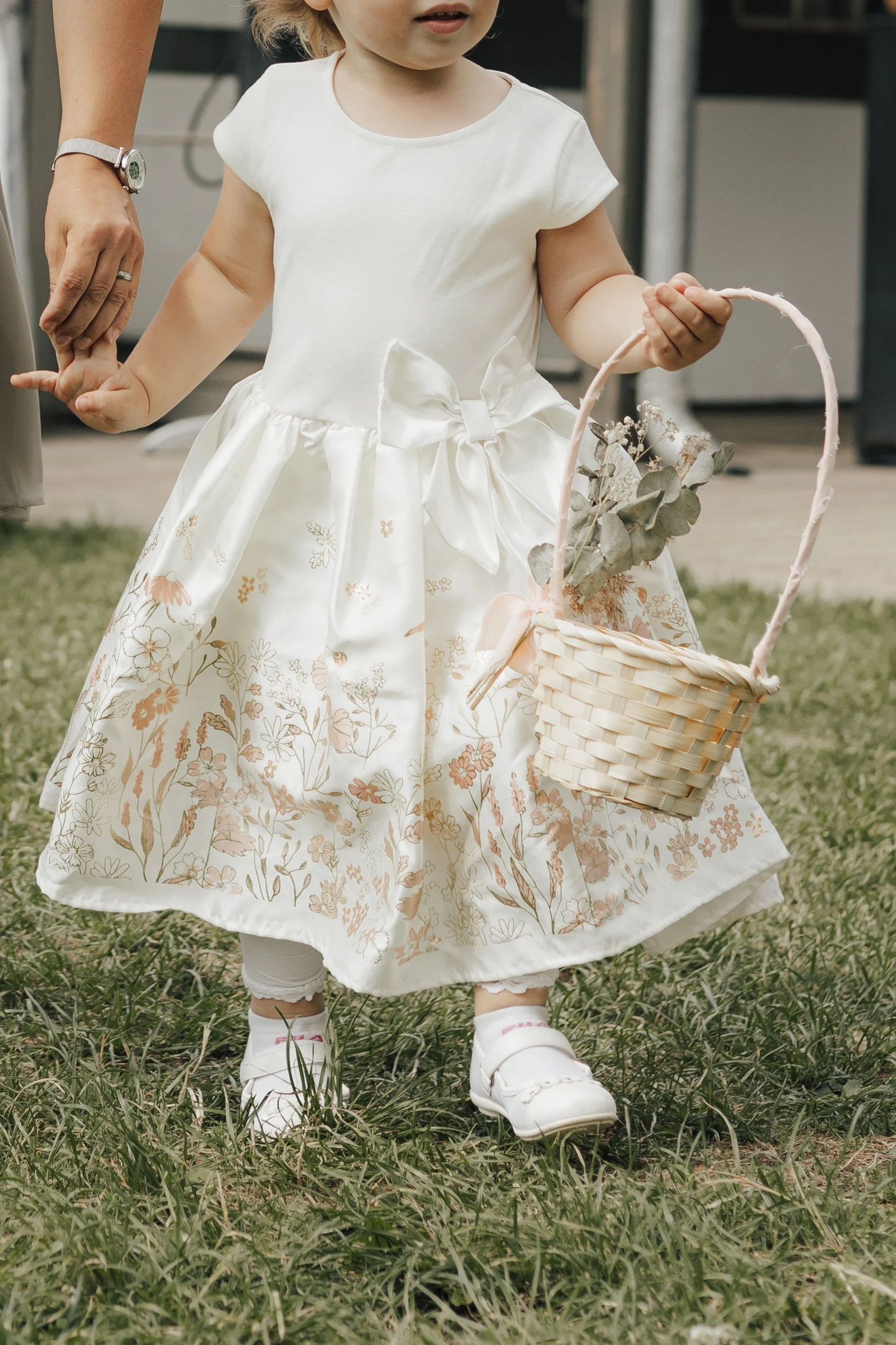 Ein kleines Mädchen trägt ein weißes Kleid mit Blumenmuster und eine Schleife, steht im Gras und hält einen Korb mit Eukalyptuszweigen. Jemand hält ihre Hand.