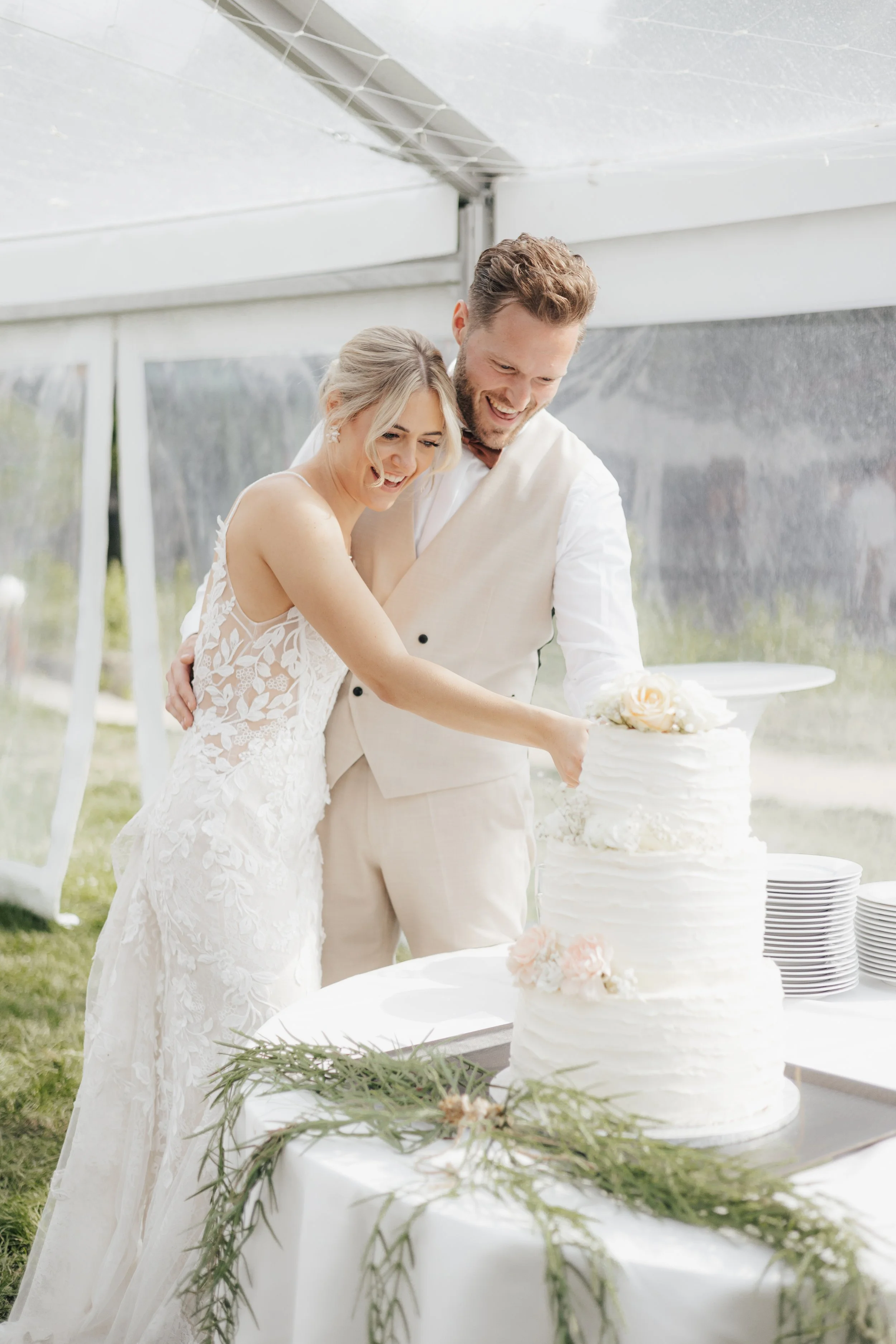Ein glückliches Brautpaar schneidet eine Hochzeitstorte bei einer freien Trauung im Freien. Das Brautkleid ist weiß mit floralem Muster, der Bräutigam trägt einen cremfarbenen Anzug. Die Torte ist weiß, geschichtet und mit Blumen dekoriert, auf einem