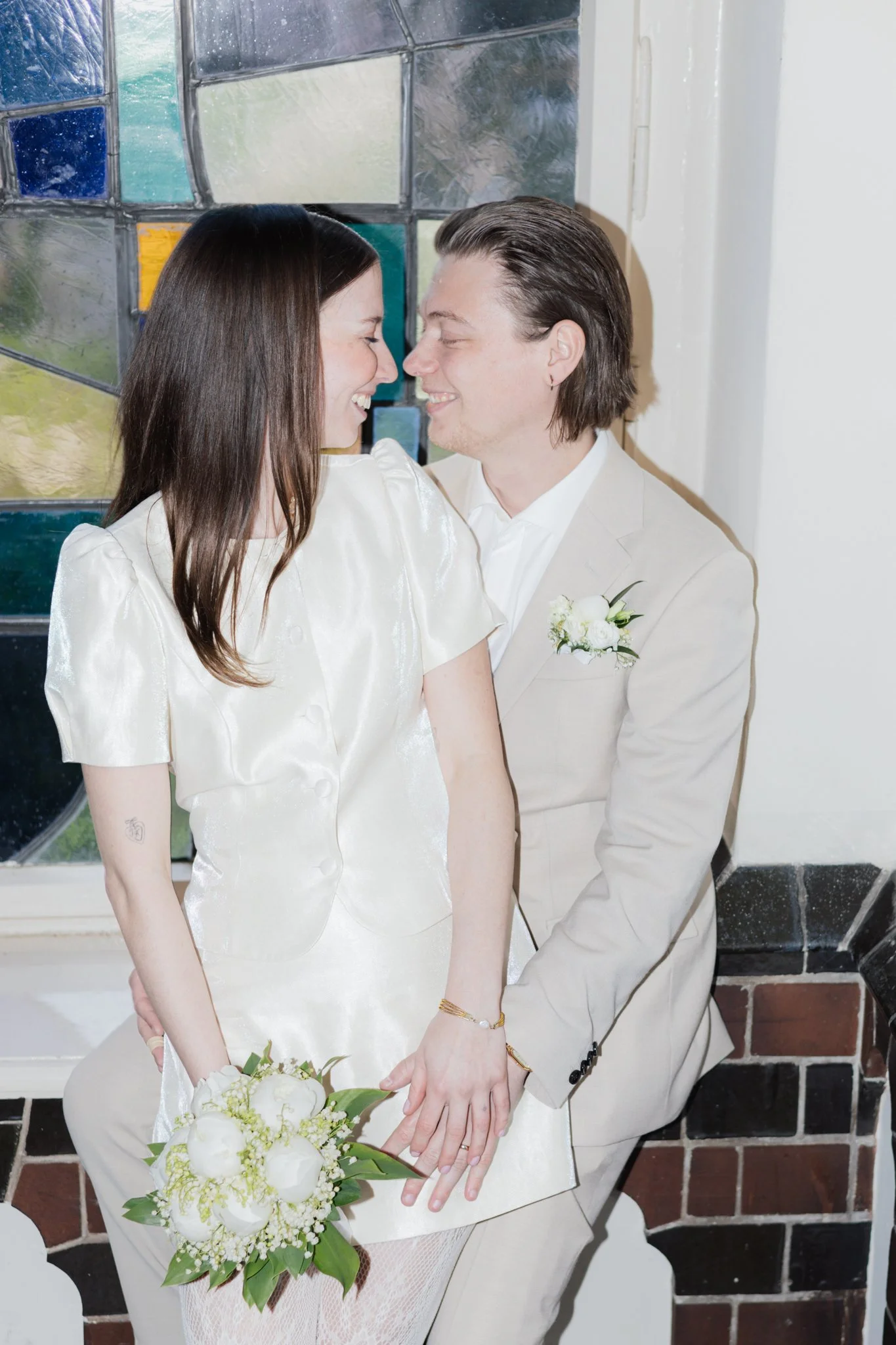 Ein Paar bei einer Hochzeit, das sich liebevoll ansieht, während sie Händchen halten und sich nahe sind, vor einer Fensterwand mit bunten Glassteinen.