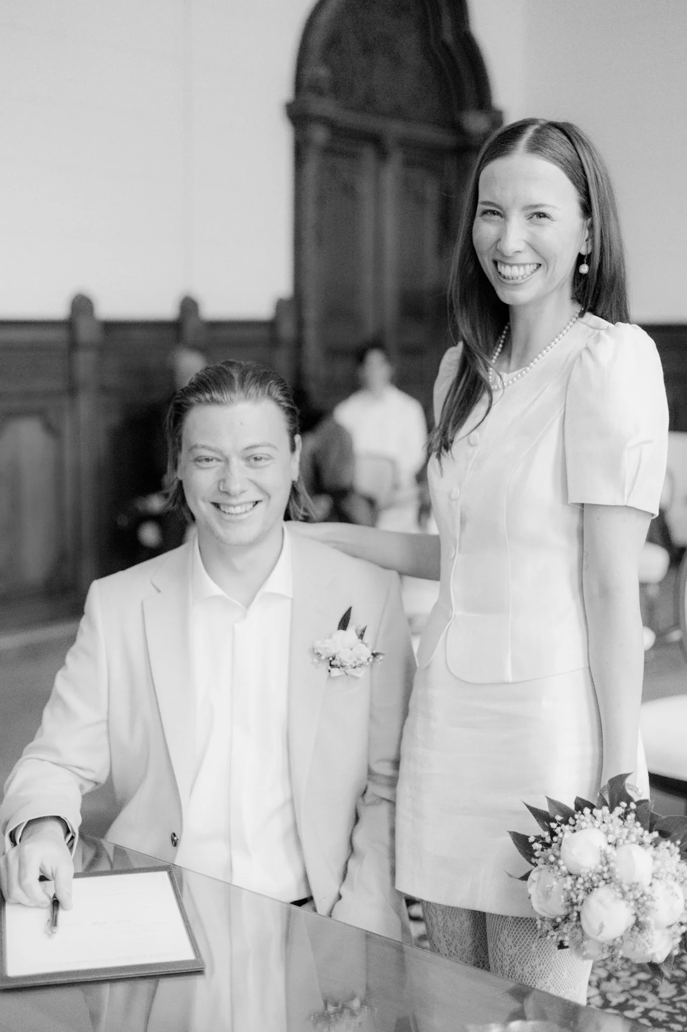 Ein Lächelndes Pärchen bei einer Hochzeit im Standesamt, der Mann unterschreibt das Dokument, die Frau hält einen Blumenstrauß, beide in formeller Kleidung.