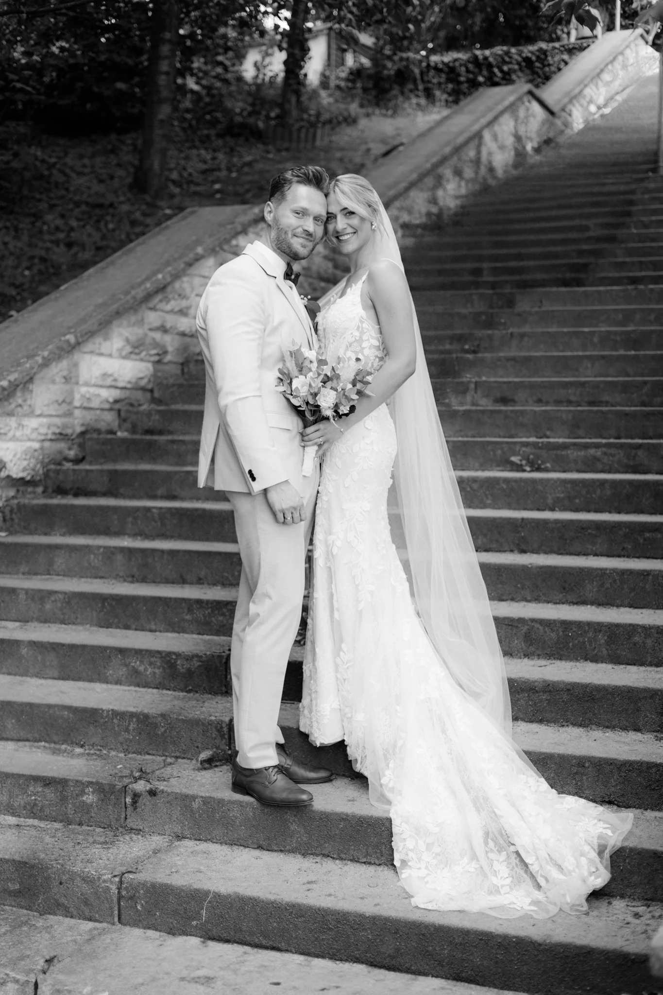 Ein lachendes Brautpaar steht auf einer Treppe im Freien, beide tragen einen weißen Hochzeitsanzug, die Braut hält einen Blumenstrauß, im Hintergrund Bäume und eine Mauer.
